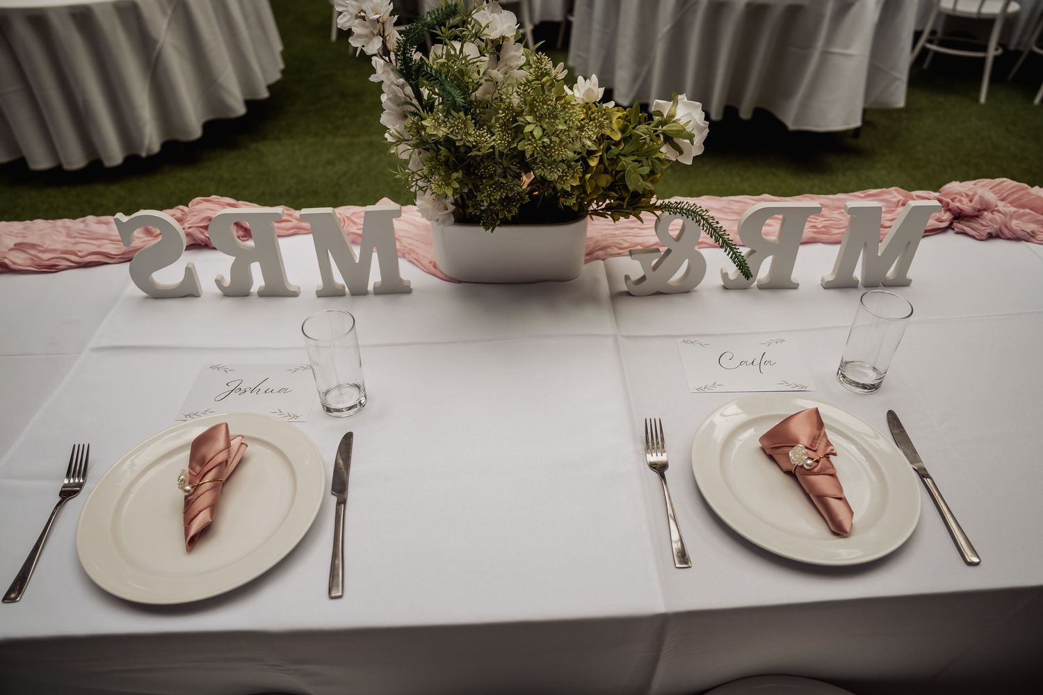 Elegant wedding head table set for bride and groom with pink napkins, floral centerpiece, and decorative MR & MRS signs.