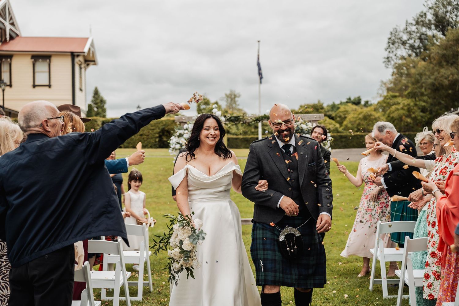Bride and groom walk down outdoor aisle as guests throw confetti at Scottish wedding ceremony.