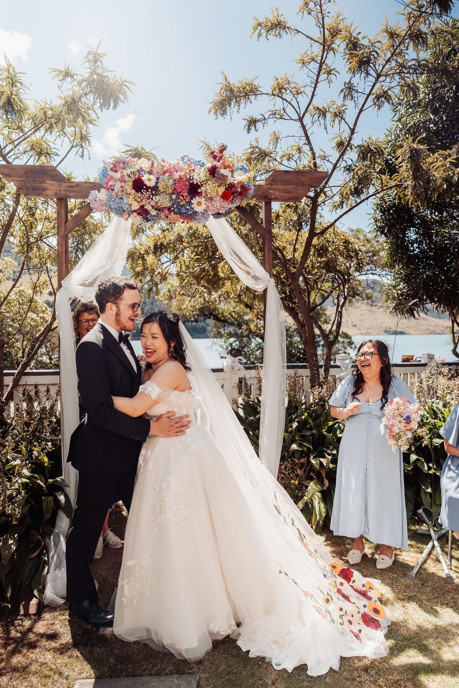 Bride and groom embrace under floral arch at outdoor wedding ceremony with bridesmaid holding bouquet.