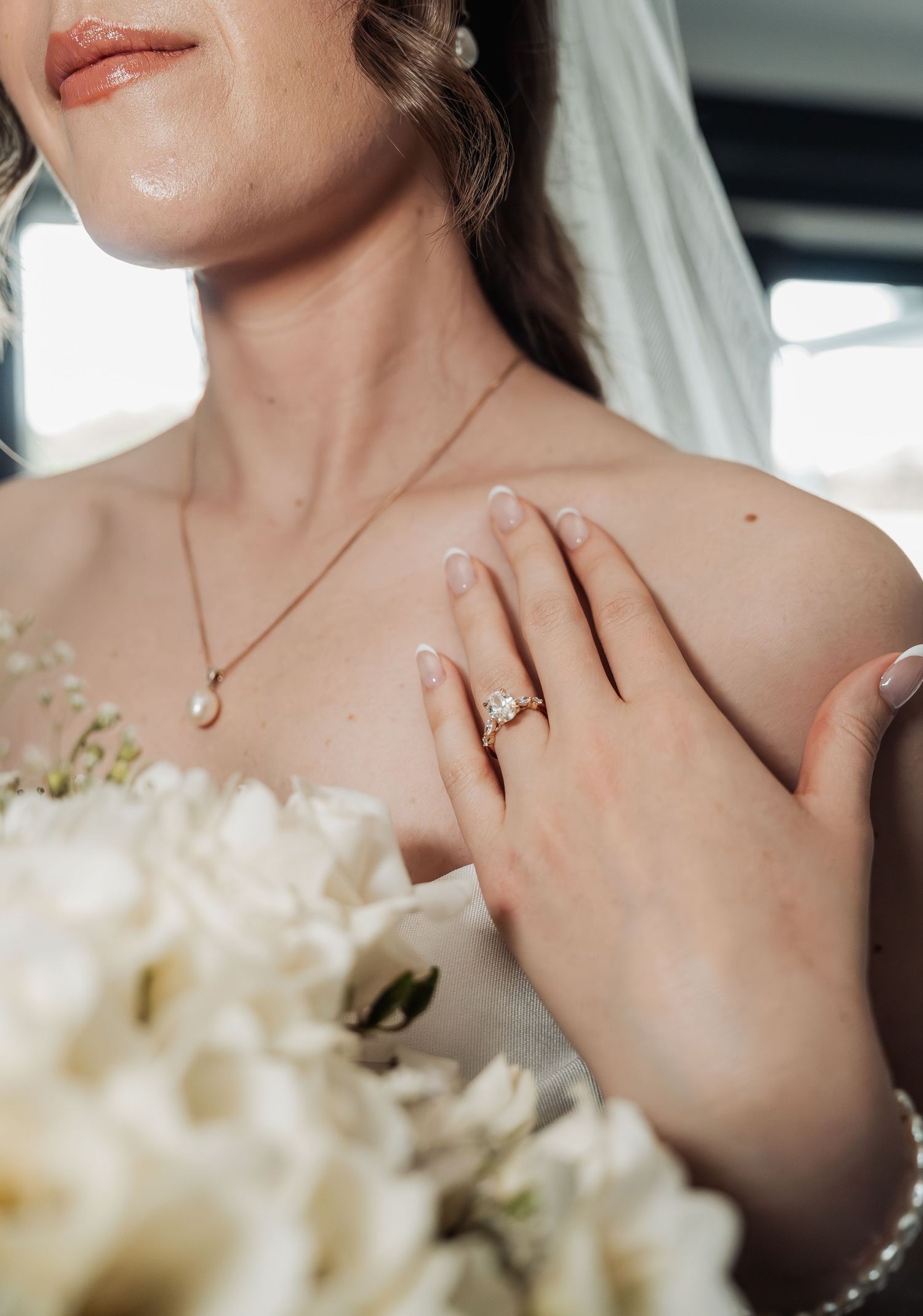 Close-up of a bride showcasing her diamond engagement ring and pearl necklace while holding a white floral bouquet.