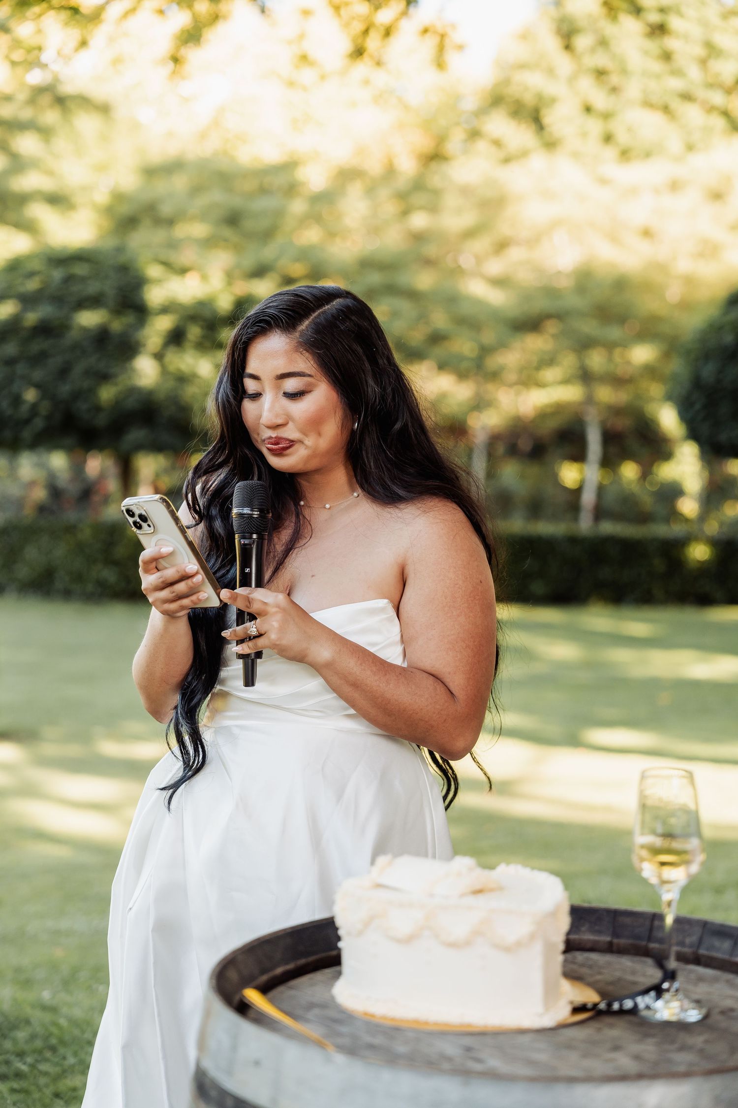 Bride in strapless white gown holds microphone and phone near wedding cake outdoors.