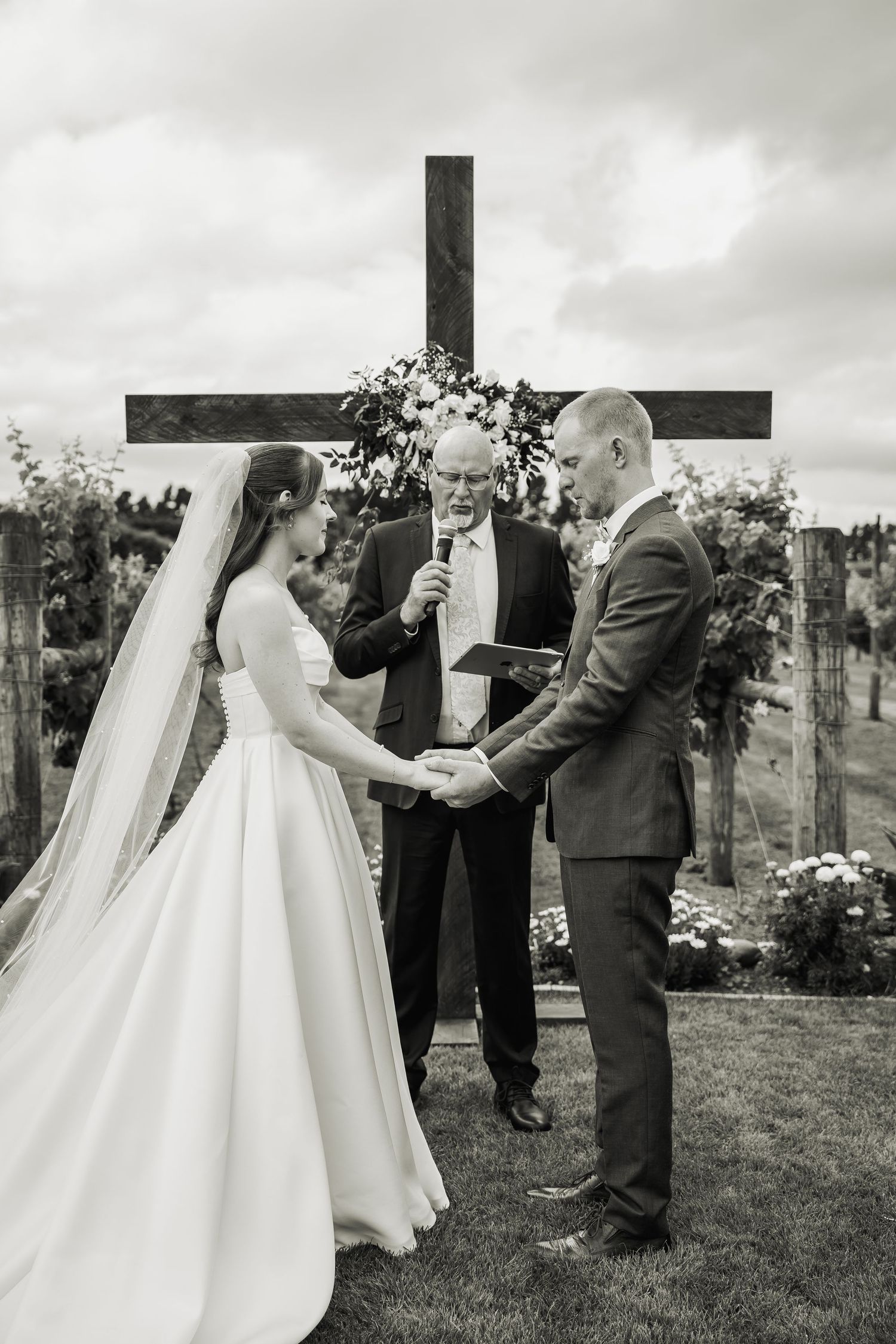 Black and white photo of bride and groom holding hands during outdoor vineyard wedding ceremony with officiant.