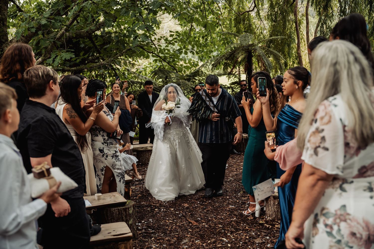 Bride in white gown walks down forest wedding aisle surrounded by guests taking photos.