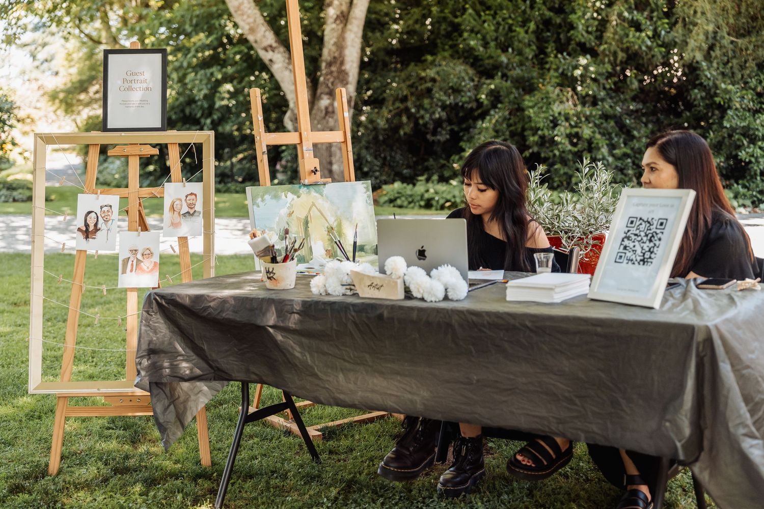 Two women sit at an outdoor event registration table with easels displaying photos and a QR code sign.