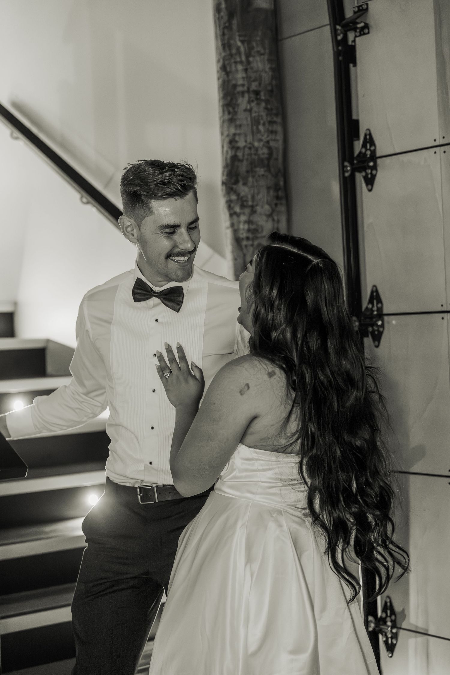 Black and white photo of smiling groom and bride near staircase with fairy lights.