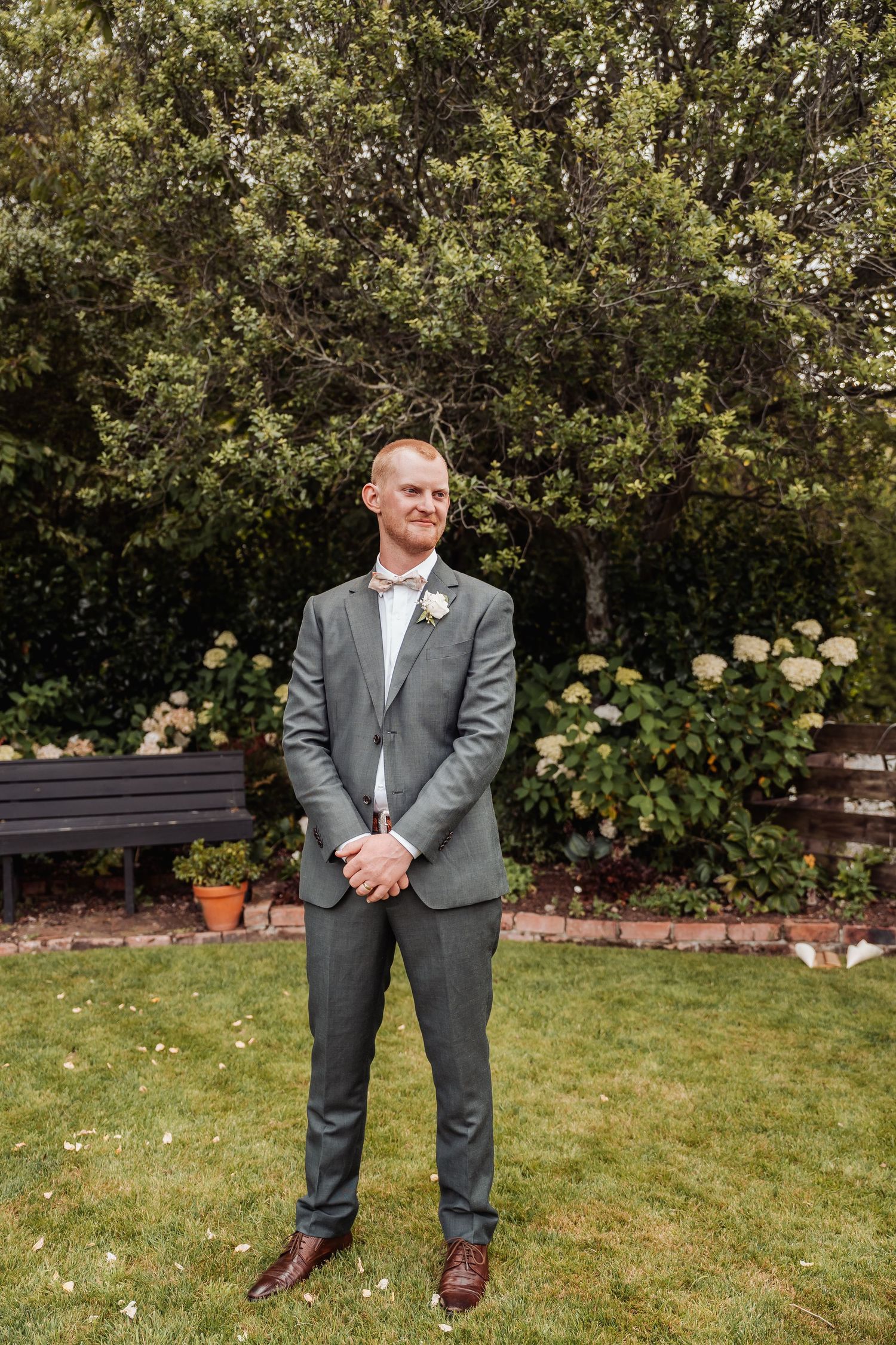 Groom in gray suit with boutonniere stands smiling on green lawn surrounded by lush garden and hydrangeas.