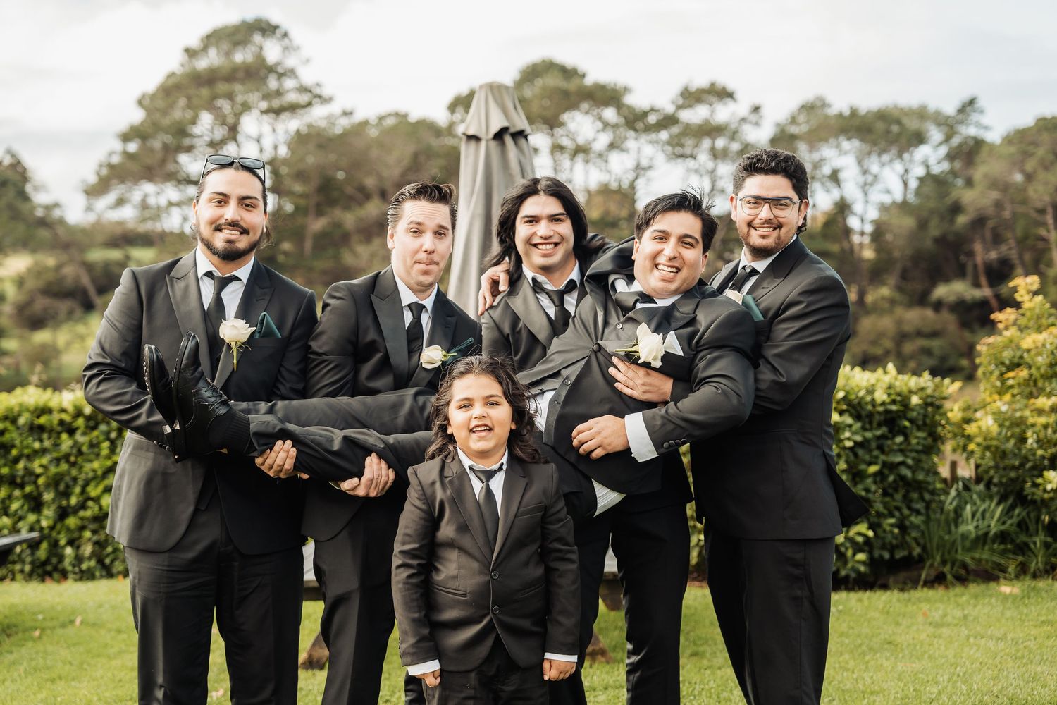 Wedding party in black suits and dresses pose together on a sunny day outdoors among green trees and grass.