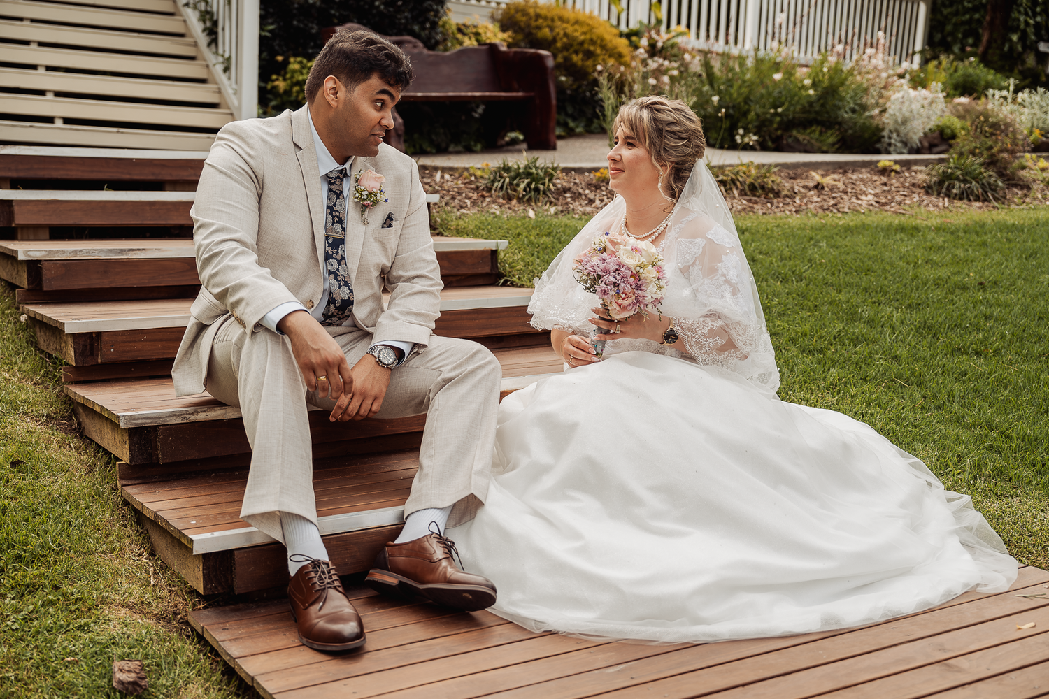Couple in wedding attire sits together on wooden steps in garden setting.