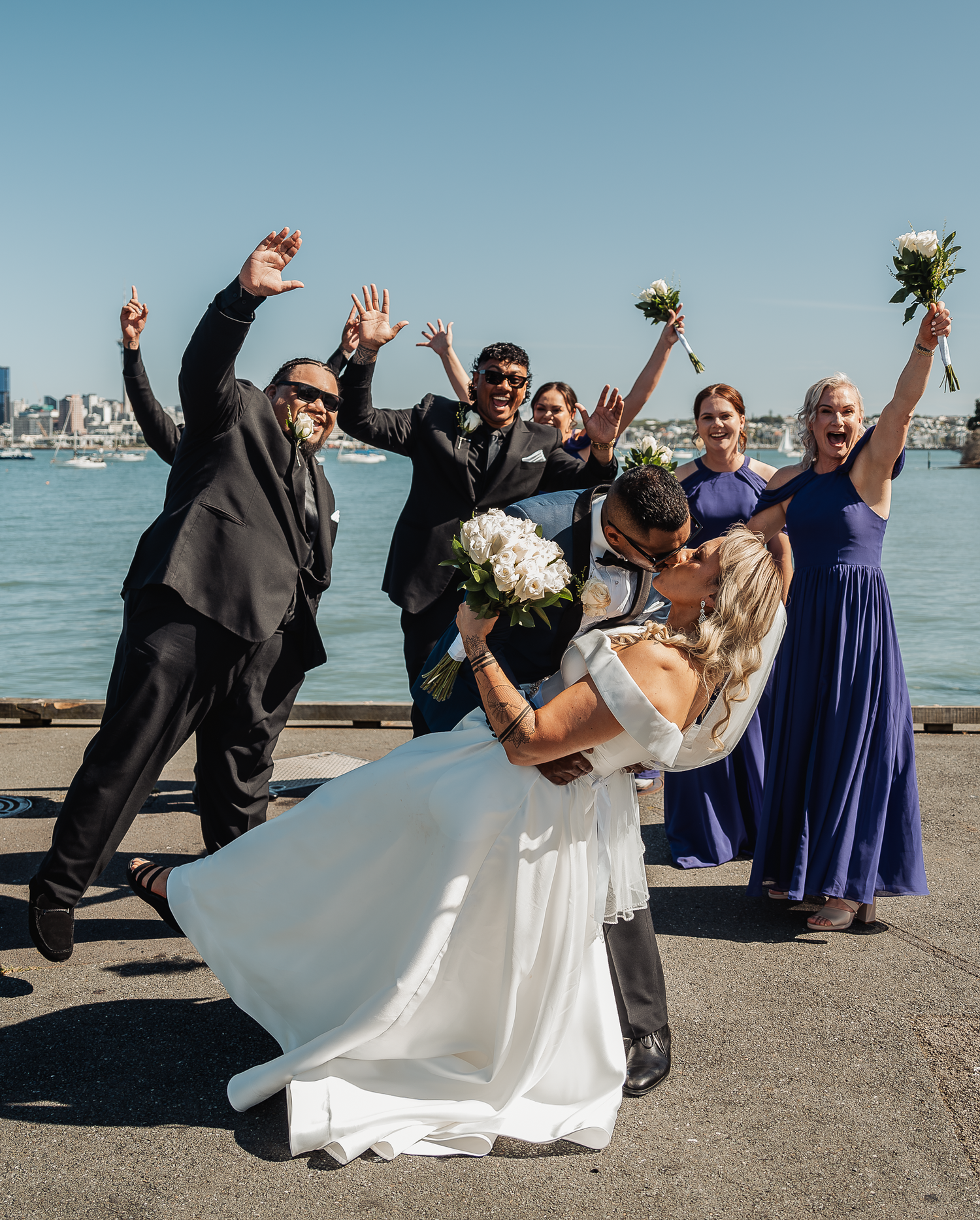 Wedding party celebrating joyfully on a waterfront pier with the city skyline in the background.