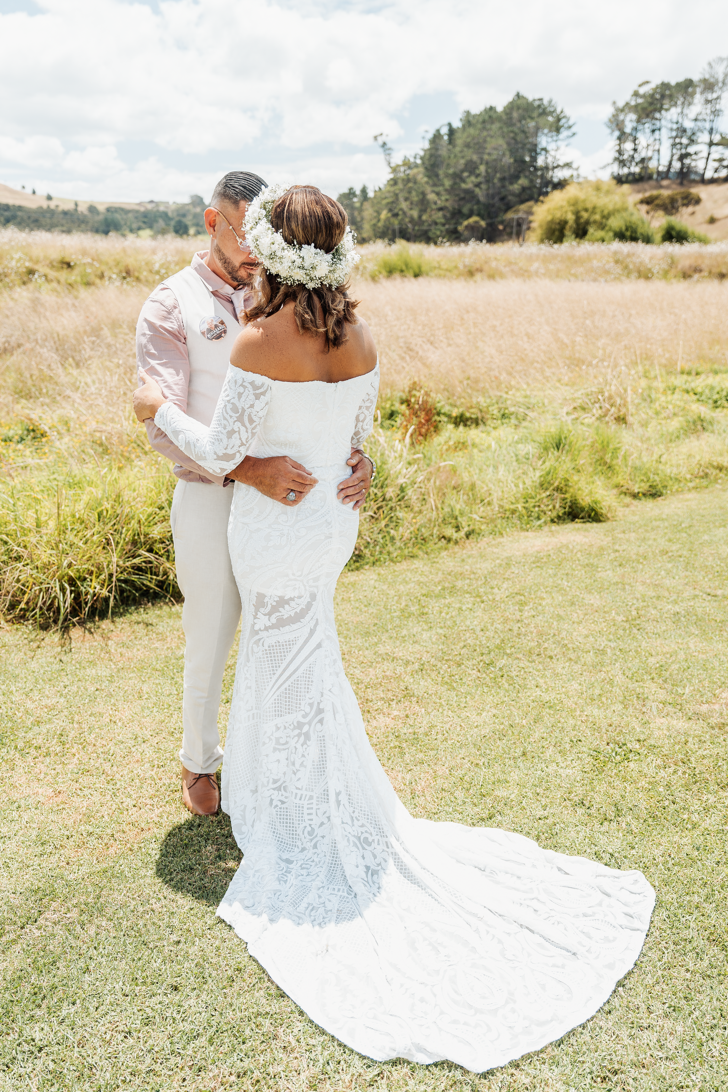 A romantic wedding photo of a couple embracing in a sunlit meadow with a flowing lace dress train.