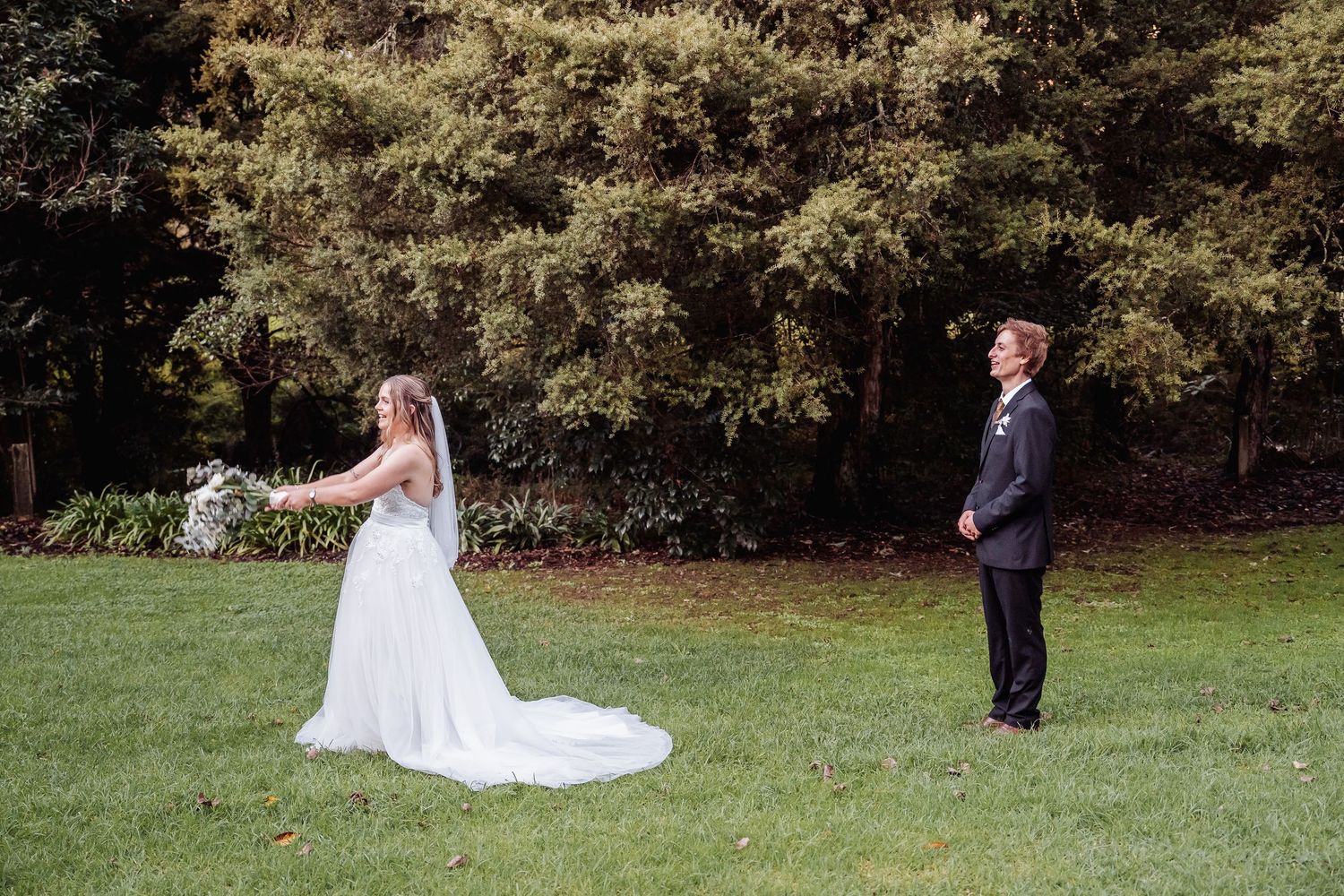 A wedding couple stands apart on green grass with trees in the background during a first look moment.
