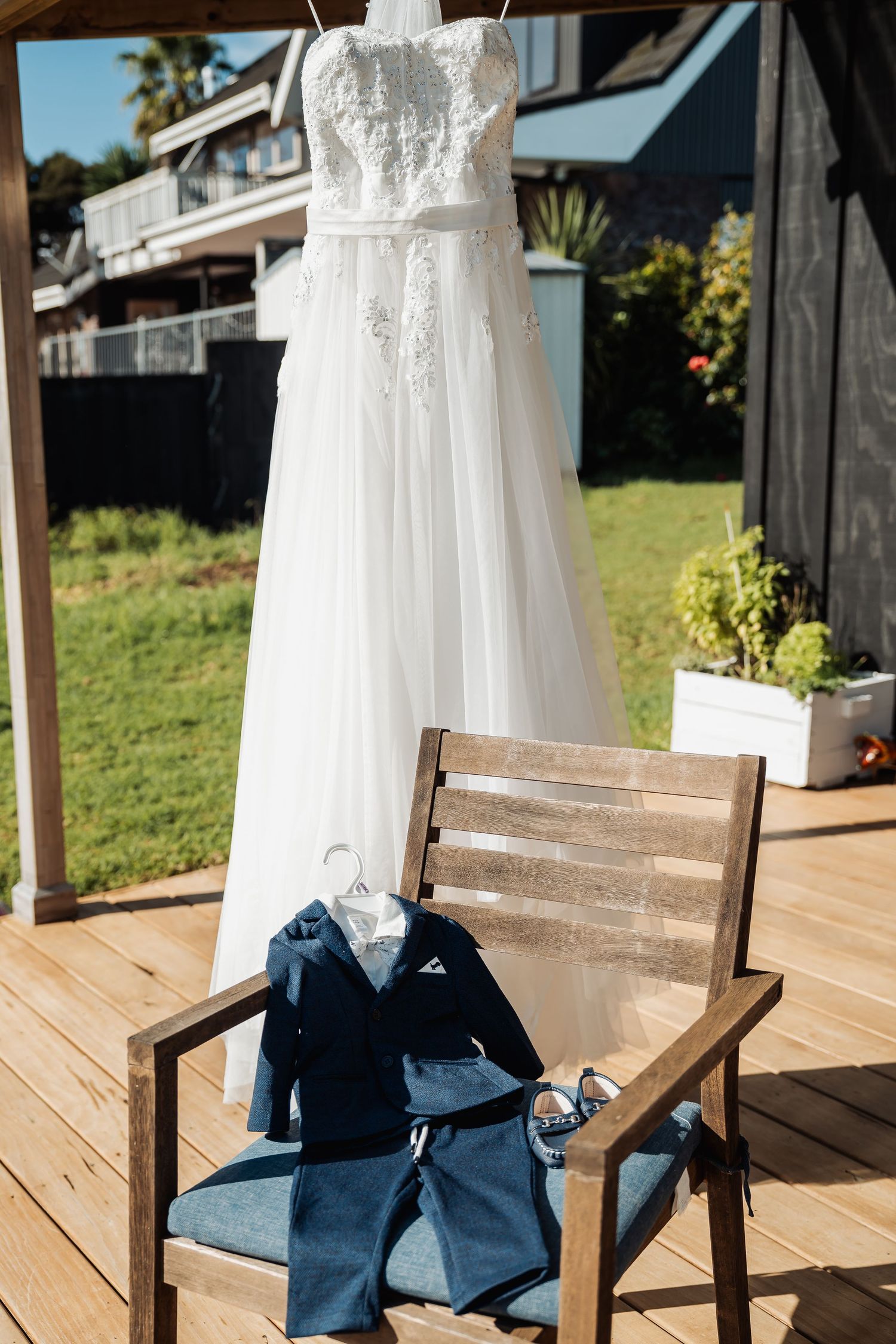 A white lace wedding dress hangs beside a wooden chair with navy blue formal attire laid out on an outdoor deck.
