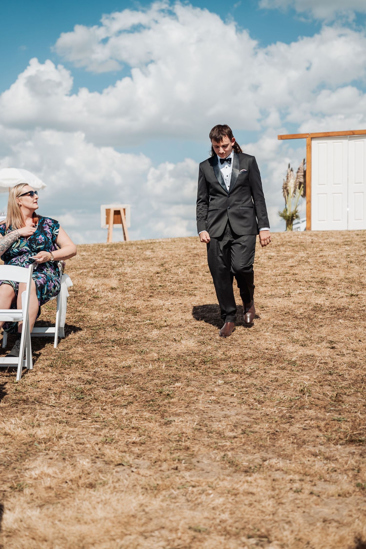 Groom in black tuxedo walks down outdoor aisle past seated guest on sunny day with clouds.