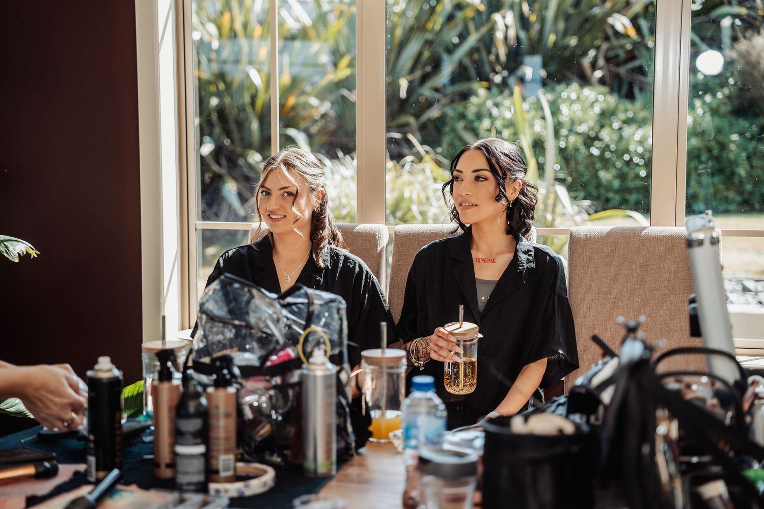 Two women in robes sit at a beauty station with hair products, smiling during bridal preparation.