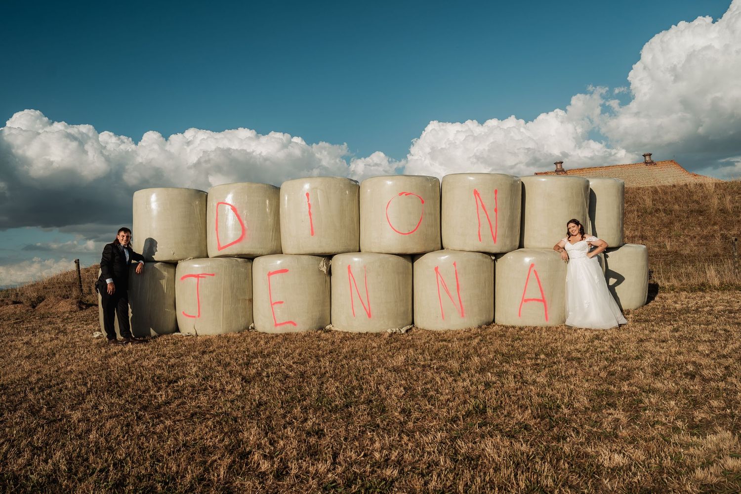 Bride and groom posing with hay bales painted with names Dion and Jenna in a rural field.