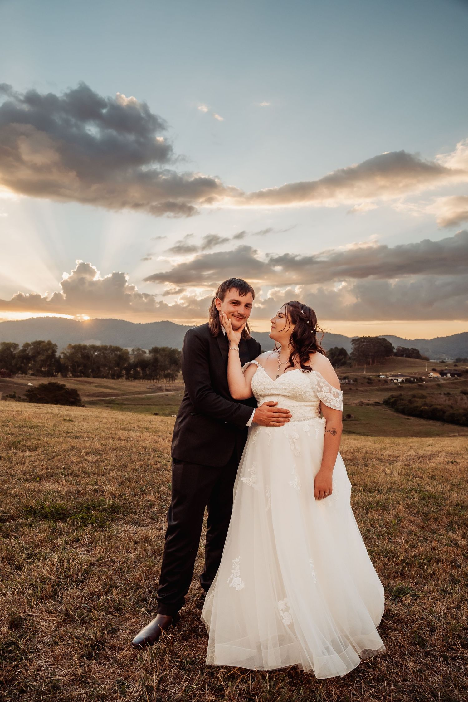 Bride and groom embracing in a golden field at sunset with rolling hills and dramatic rays of light behind them.