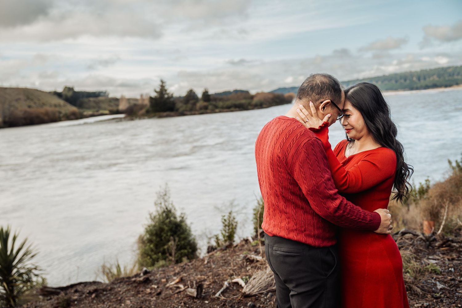 Romantic couple in red embracing forehead-to-forehead near a scenic river on an overcast day.