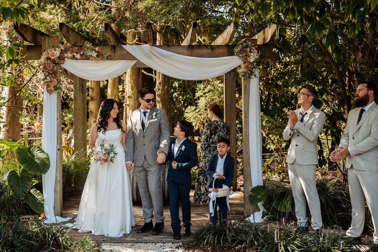 Bride and groom stand with children under a draped garden arbor during an outdoor wedding ceremony.