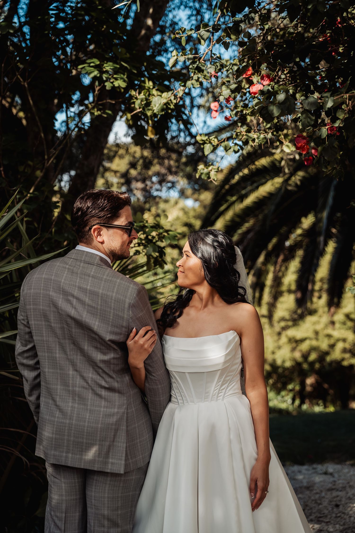 Bride and groom share a romantic moment surrounded by lush green trees and red flowering branches.