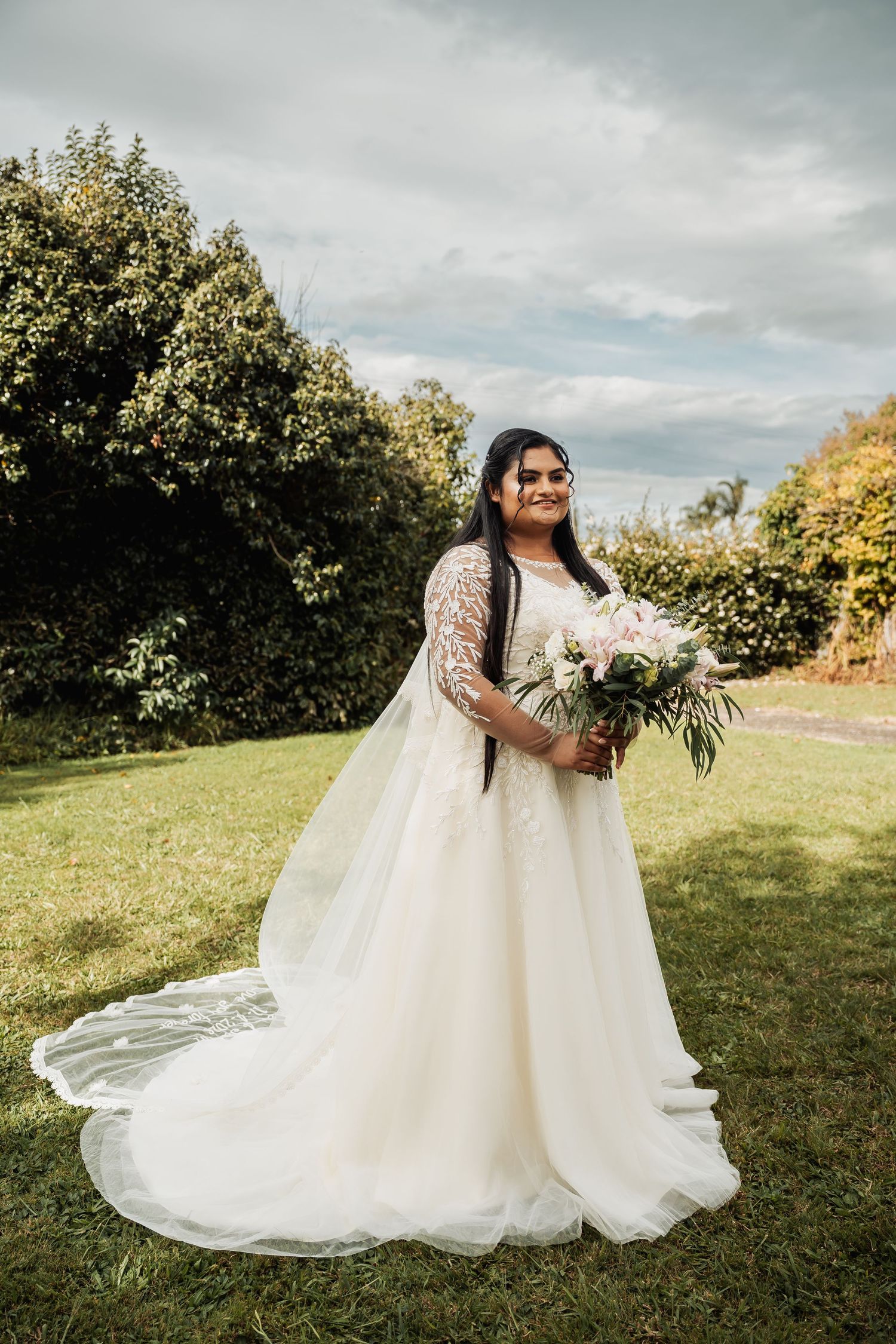 Bride in lace wedding gown holding bouquet of pink and white flowers posing in garden setting.