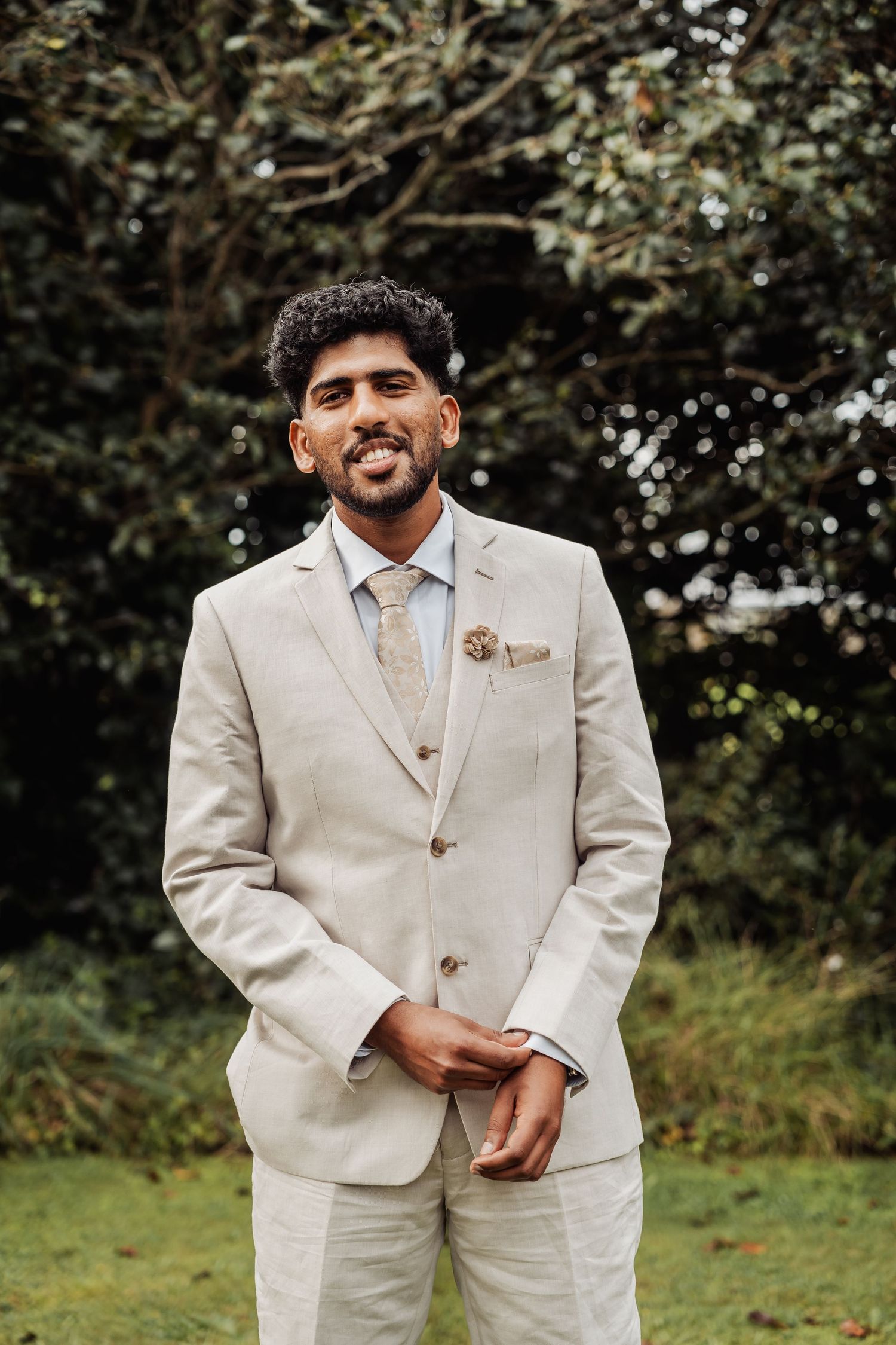 Groom in beige linen suit smiling outdoors with lush green trees in background.
