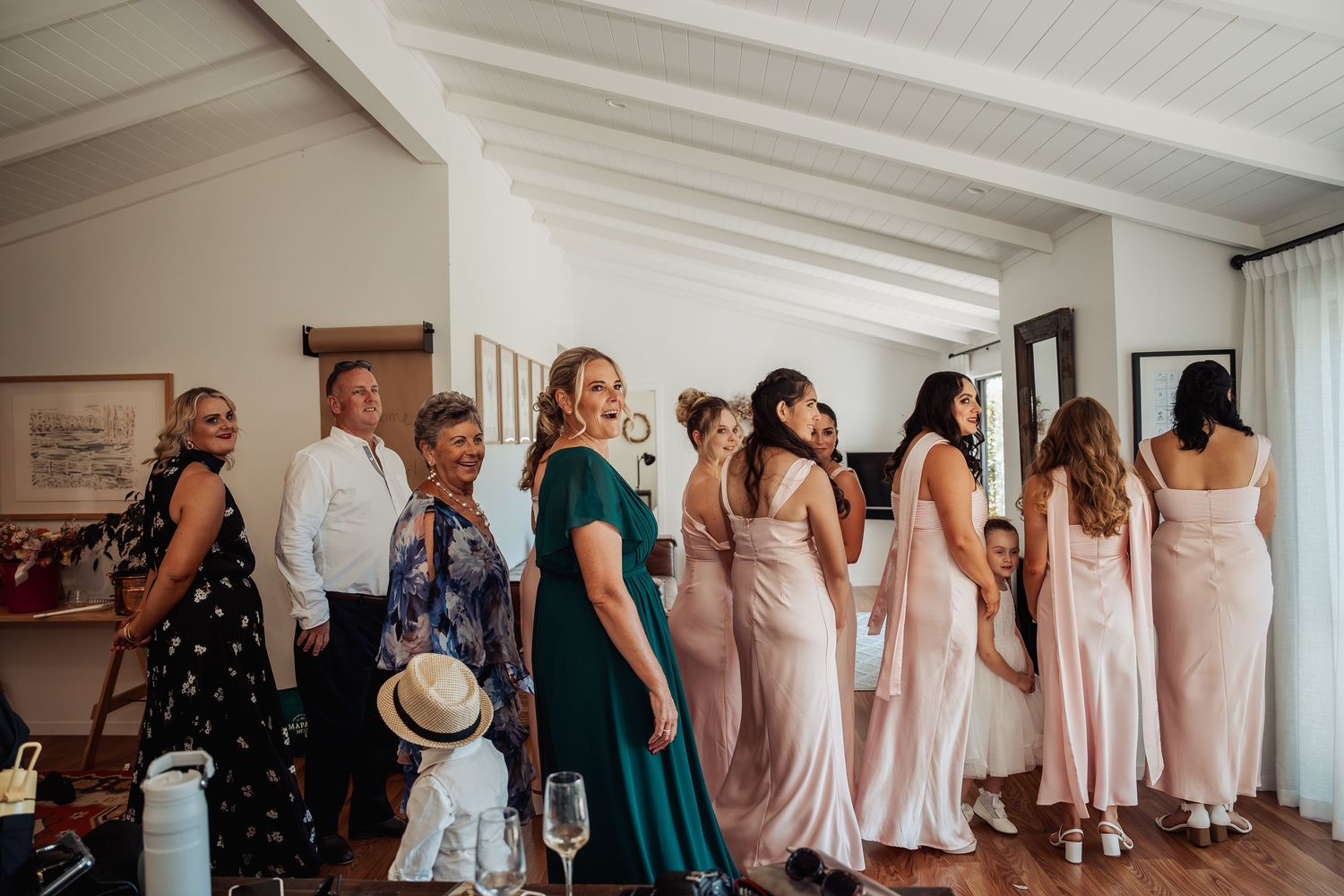 Bridesmaids in blush pink gowns and guests gathered in a bright room during wedding preparations.