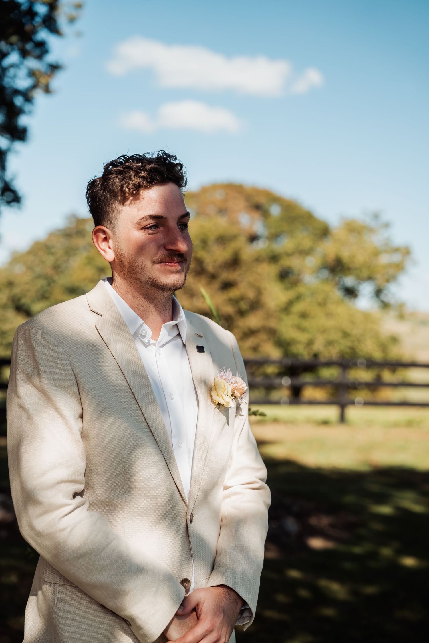 Groom in cream suit with floral boutonniere standing outdoors on a sunny wedding day.
