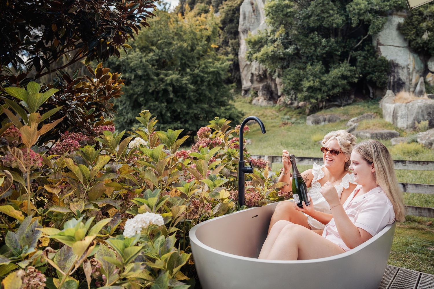 Two women relaxing in an outdoor stone bathtub surrounded by lush garden greenery.