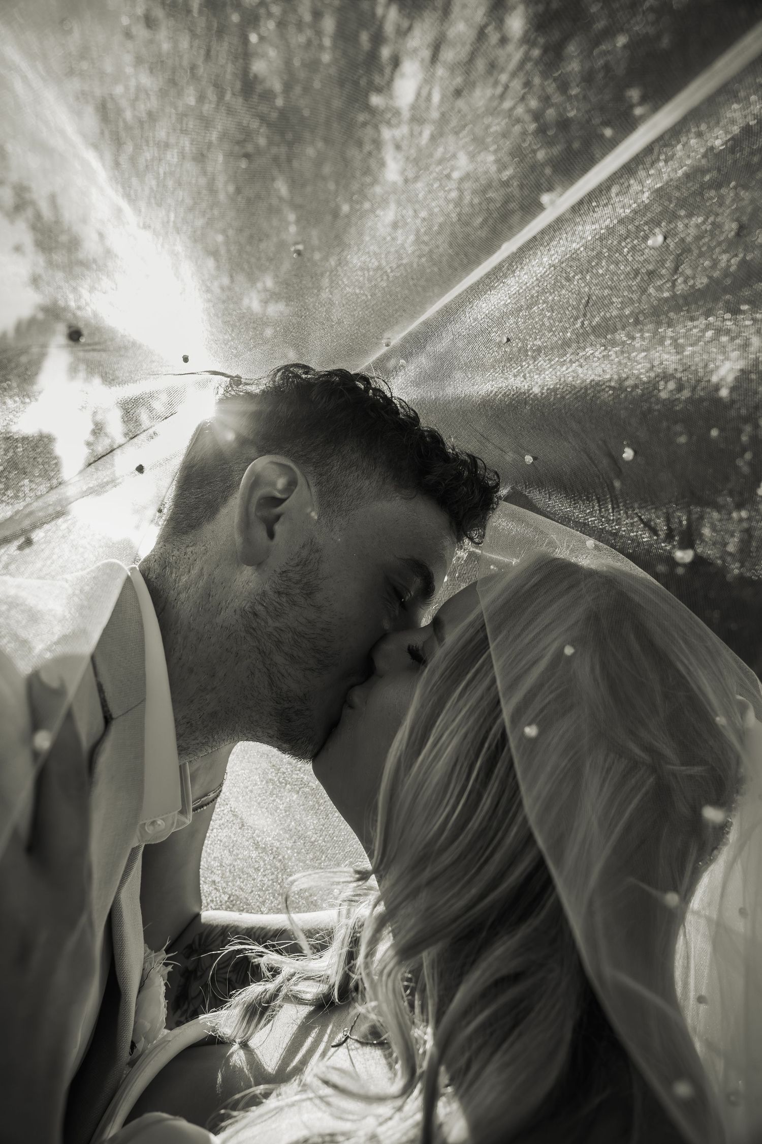 Black and white romantic photo of bride and groom kissing beneath a sparkling wedding veil.