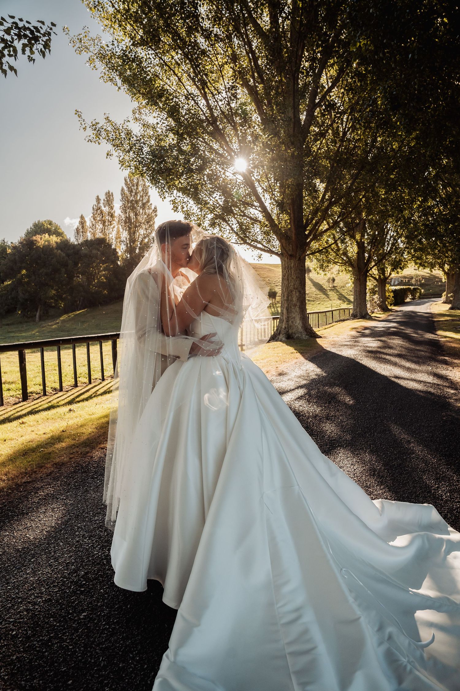Bride and groom kiss on a sunlit tree-lined path, bridal veil and gown train flowing beautifully.