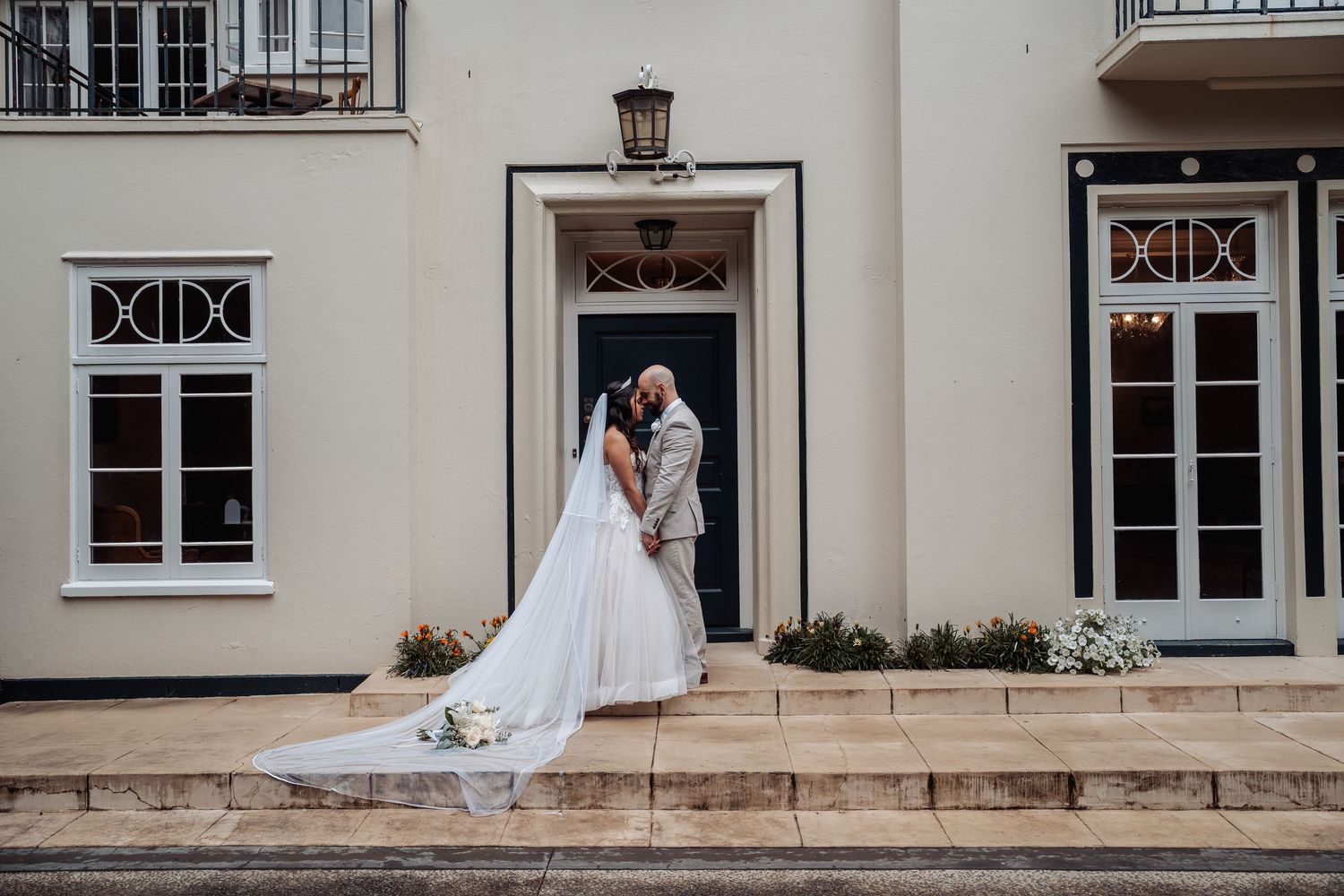 Bride and groom share a kiss on steps outside an elegant cream building with black door.
