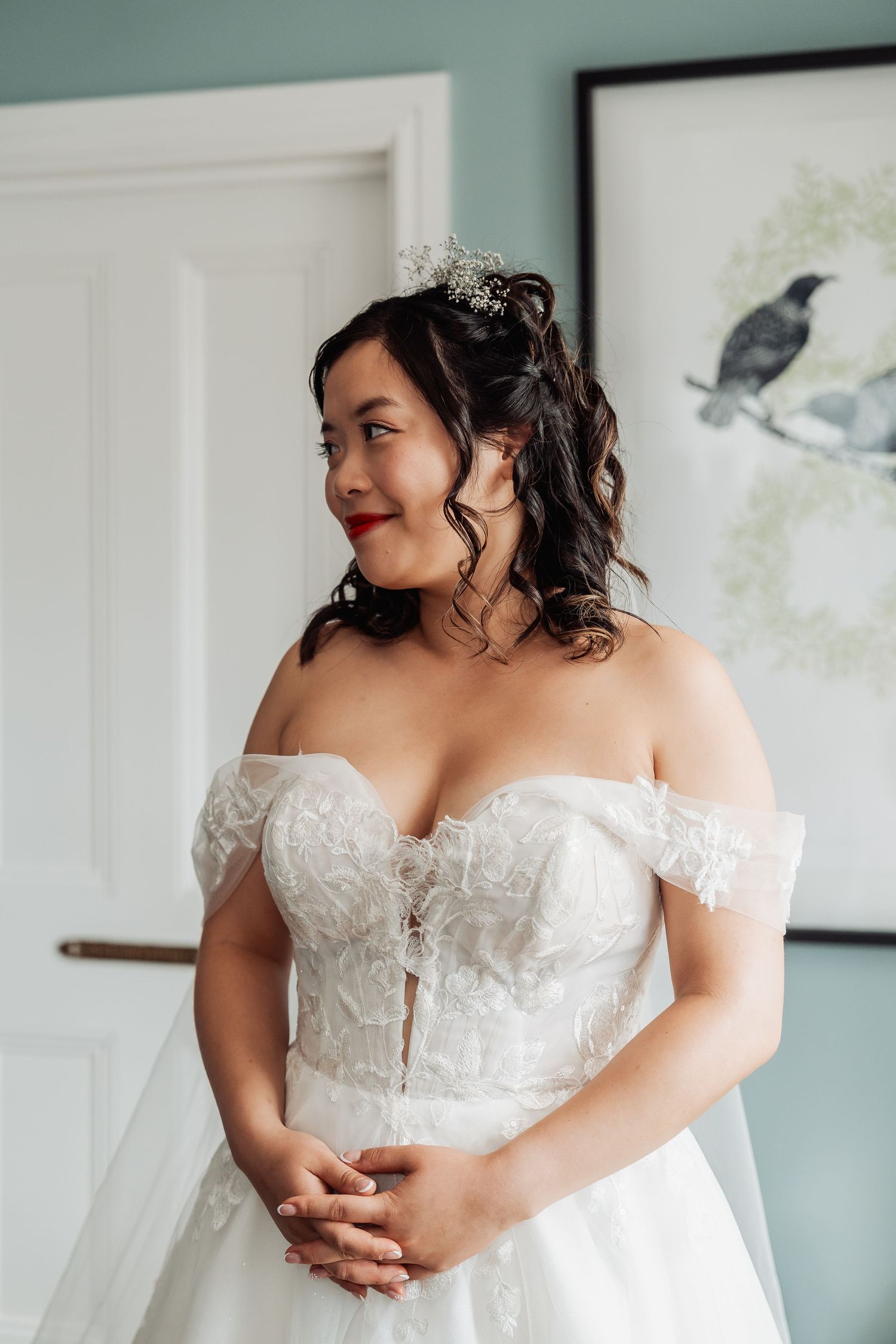 Bride in off-shoulder lace wedding gown with floral hair accessory, smiling elegantly in a softly lit room.