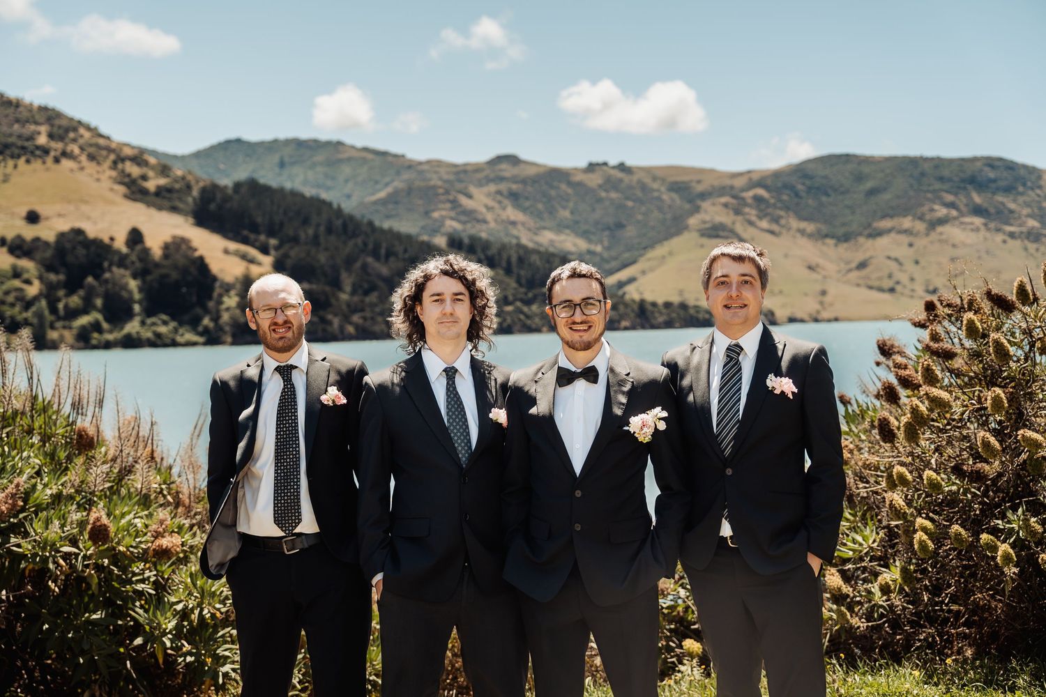 Four groomsmen in navy suits with boutonnieres pose outdoors with scenic lake and green hills backdrop.