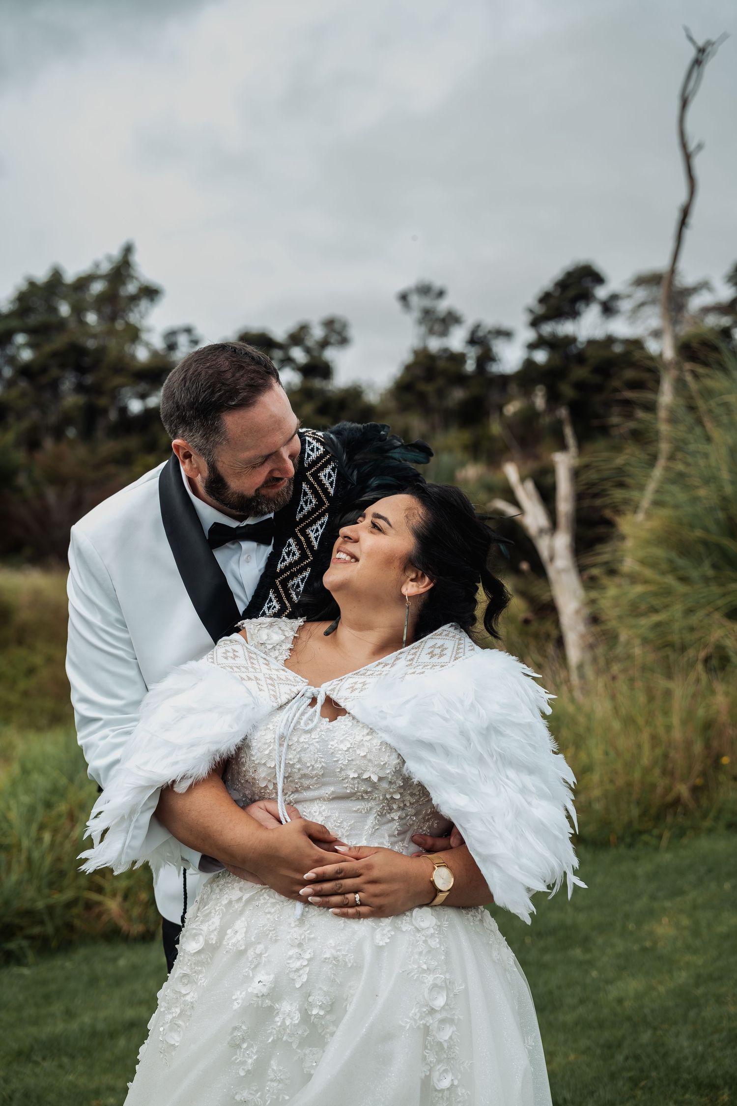 Smiling bride in feather cape embraced by groom in white tuxedo jacket outdoors on their wedding day.