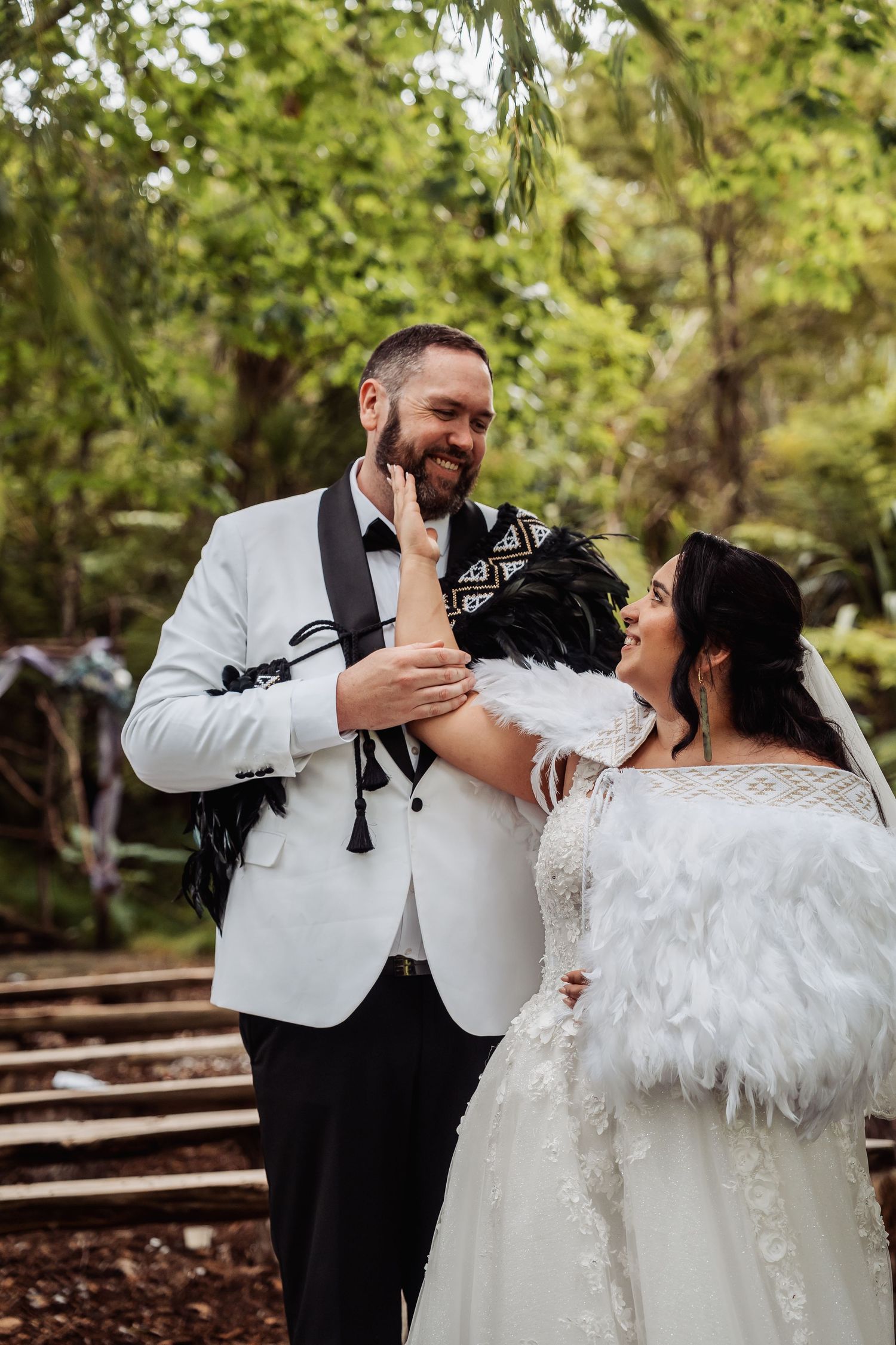 Smiling bride and groom in white attire with black feather accessories share a joyful moment in a lush green forest setting.