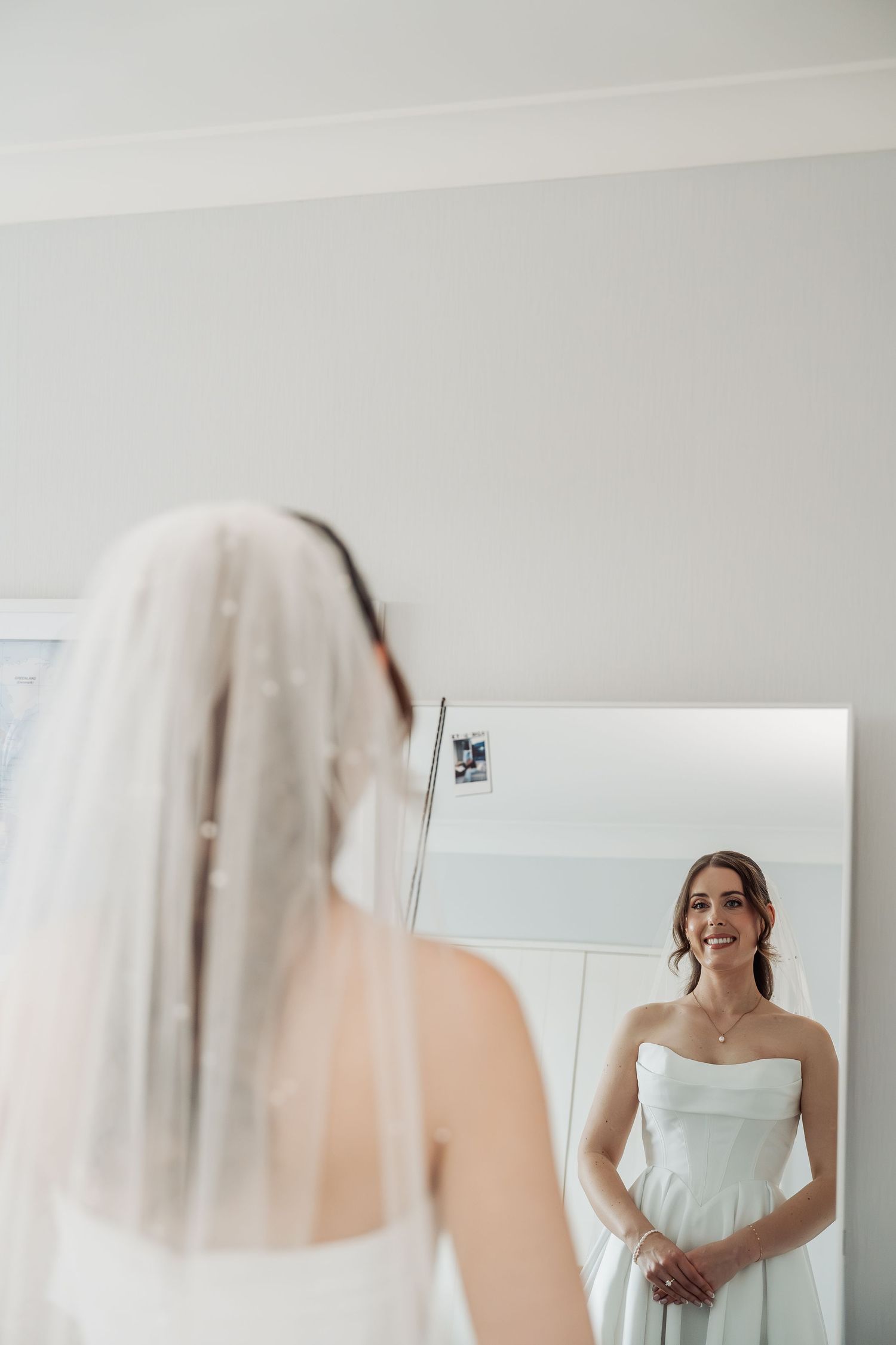 Bride in strapless white gown and veil smiling at her reflection in a full-length mirror.