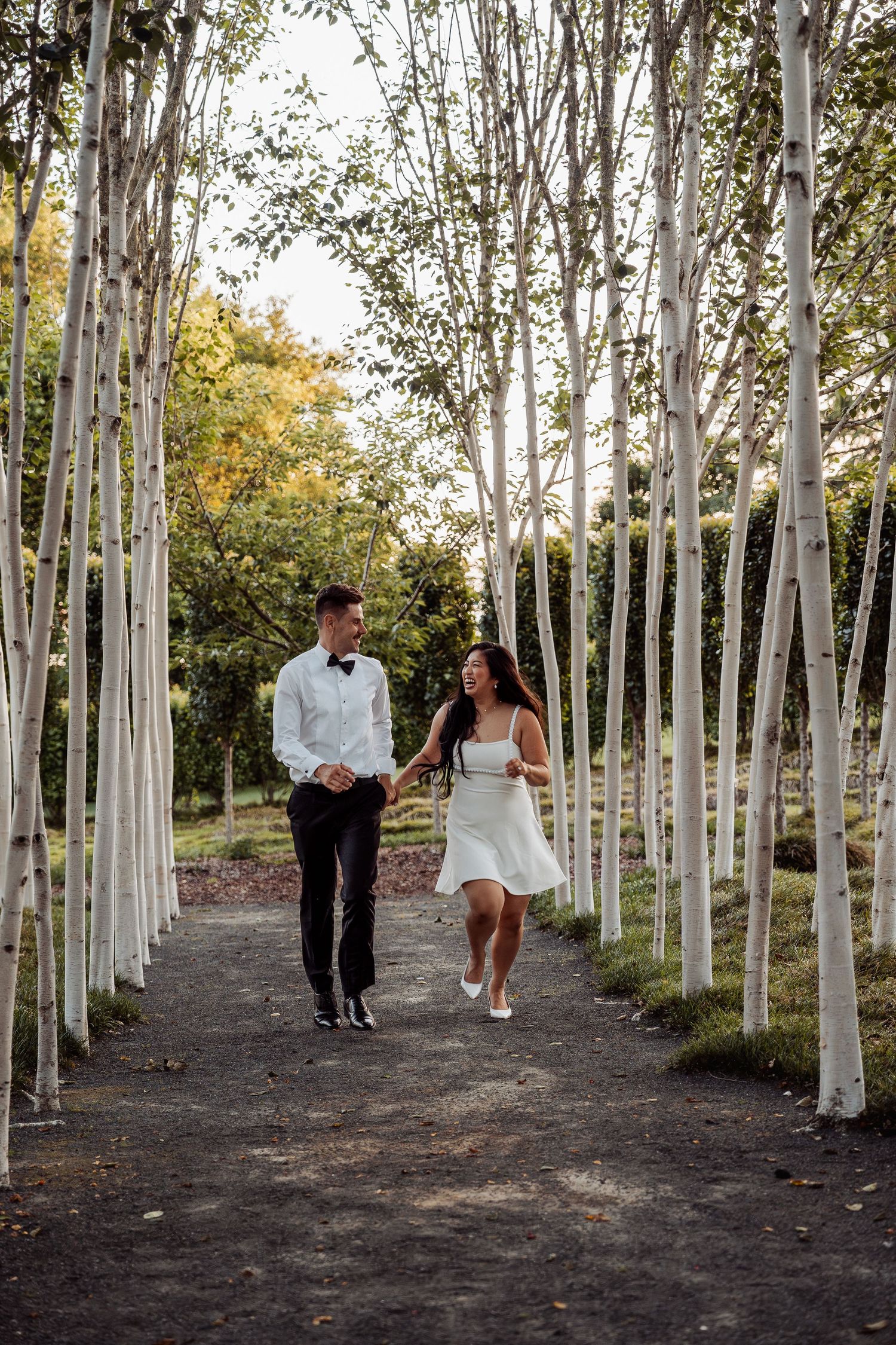 Newlyweds holding hands walking through elegant birch tree lined garden path on wedding day.
