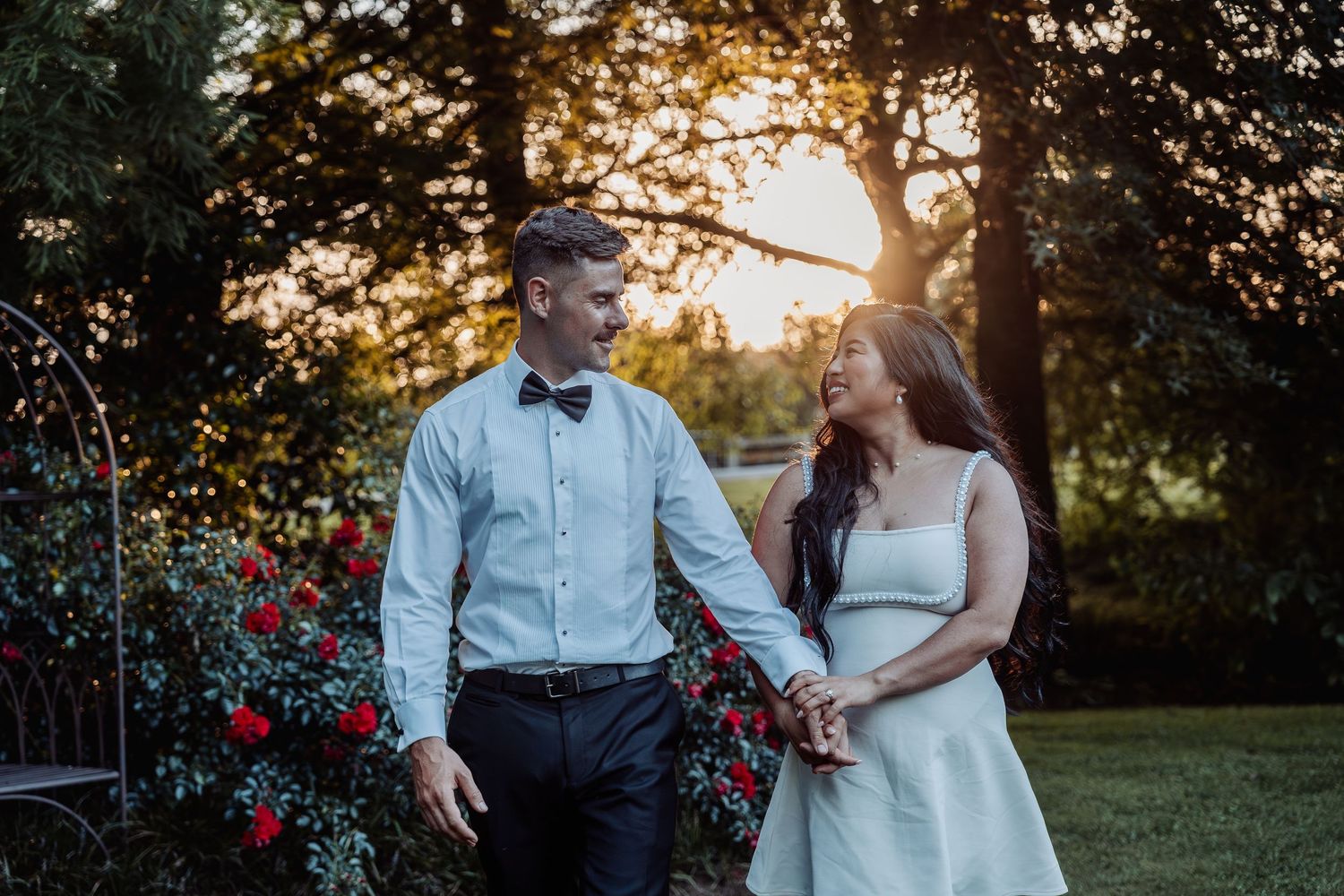 Couple in wedding attire hold hands and smile at sunset among red roses in a garden.