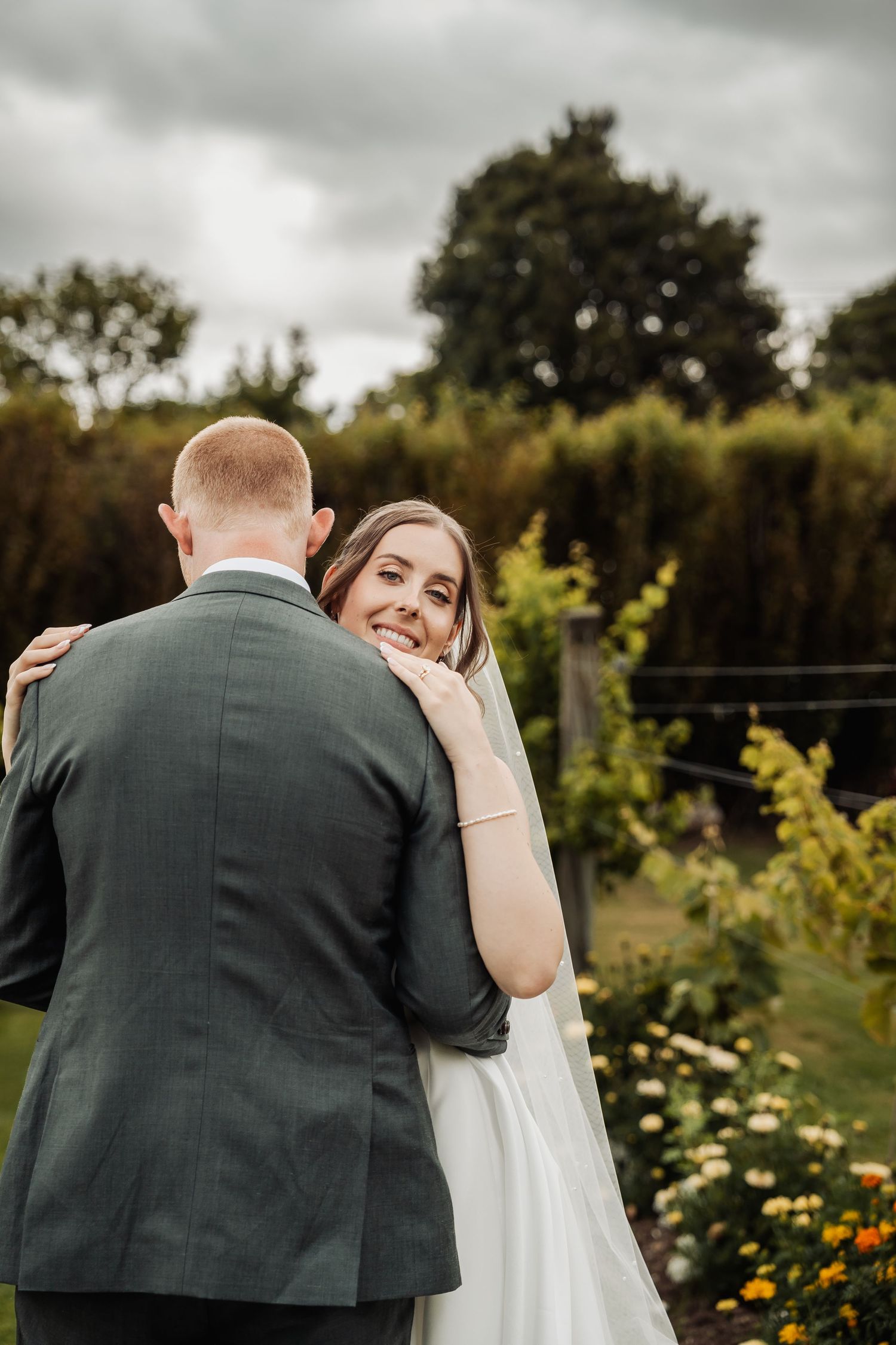 Smiling bride in white gown embraces groom in gray suit in a lush outdoor garden setting.