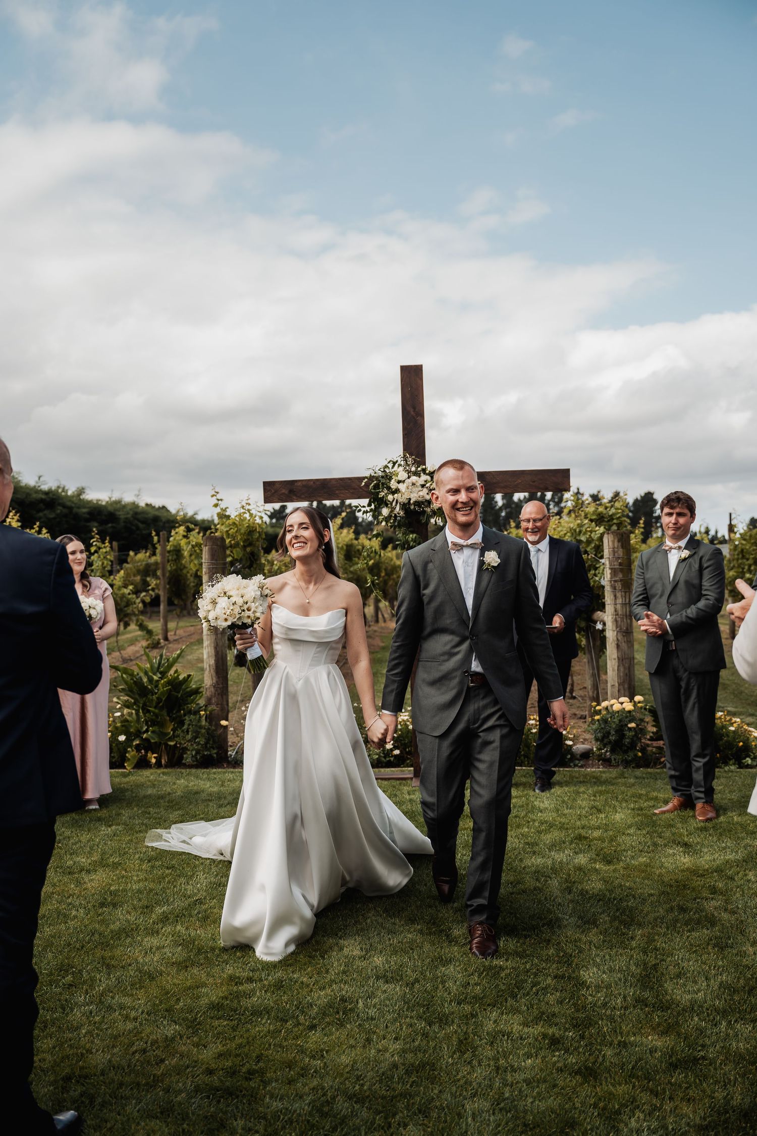Joyful newlyweds walk hand-in-hand down outdoor aisle at vineyard wedding with wooden cross backdrop.