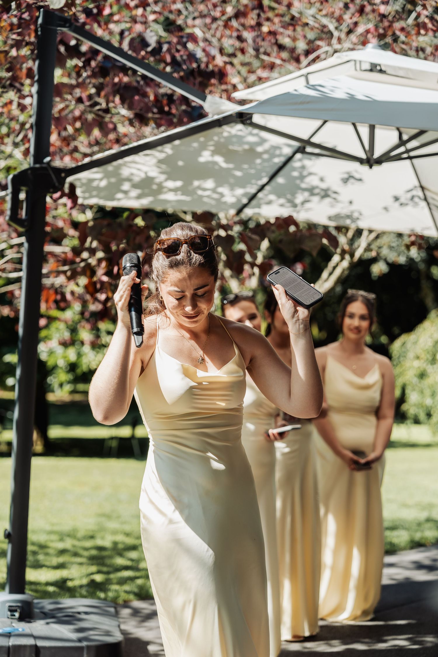 Bridesmaid in yellow satin dress holds microphone and phone while giving speech outdoors at wedding reception.