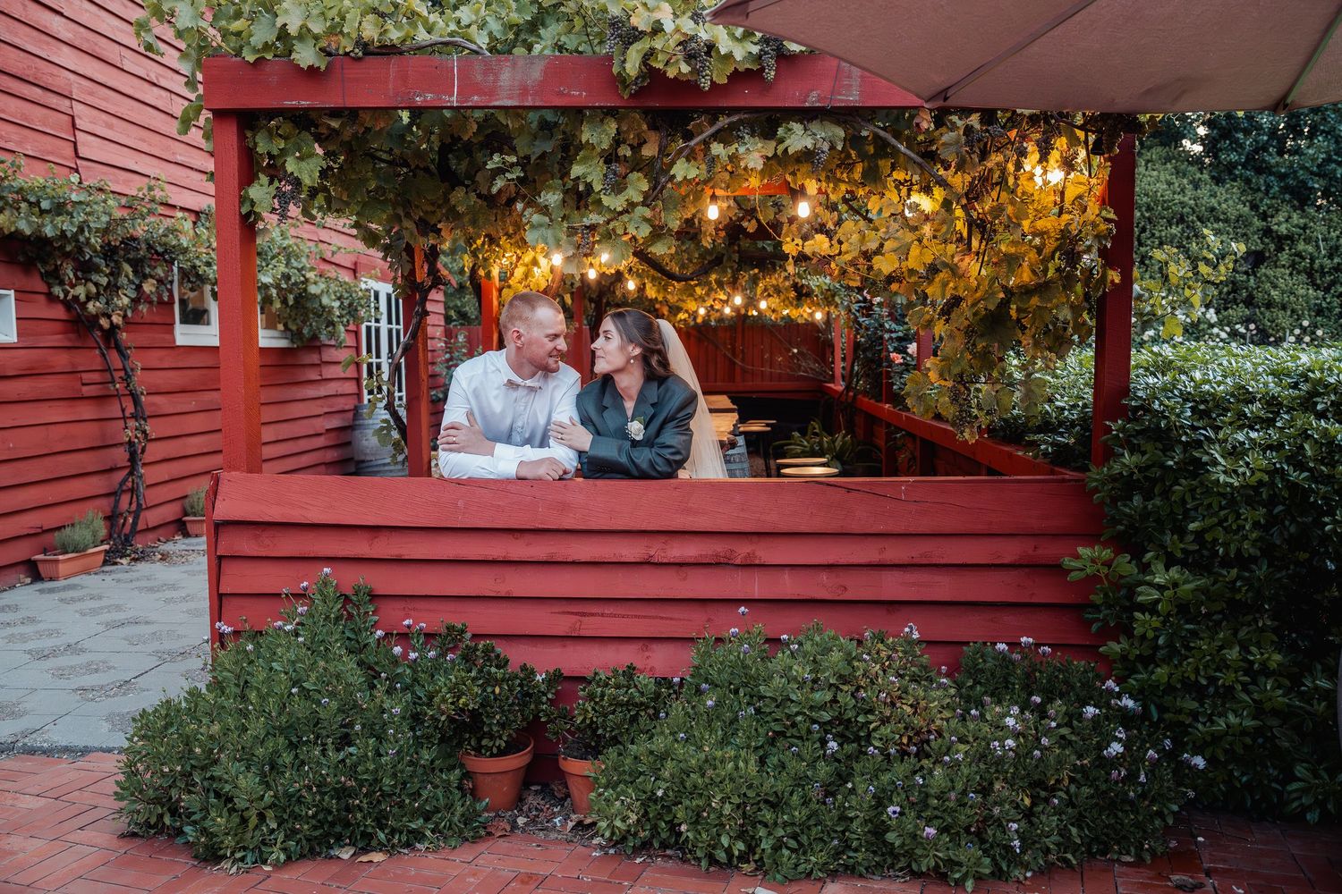 Couple sits together under a red pergola with fairy lights surrounded by lush greenery.