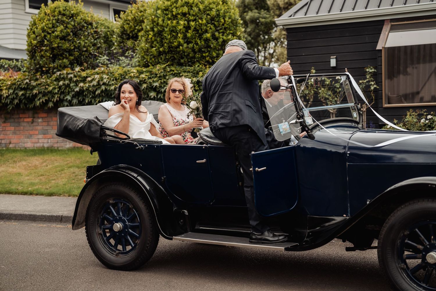 Two women ride in a vintage navy blue convertible classic car, driven by a man in a suit, on a tree-lined street.