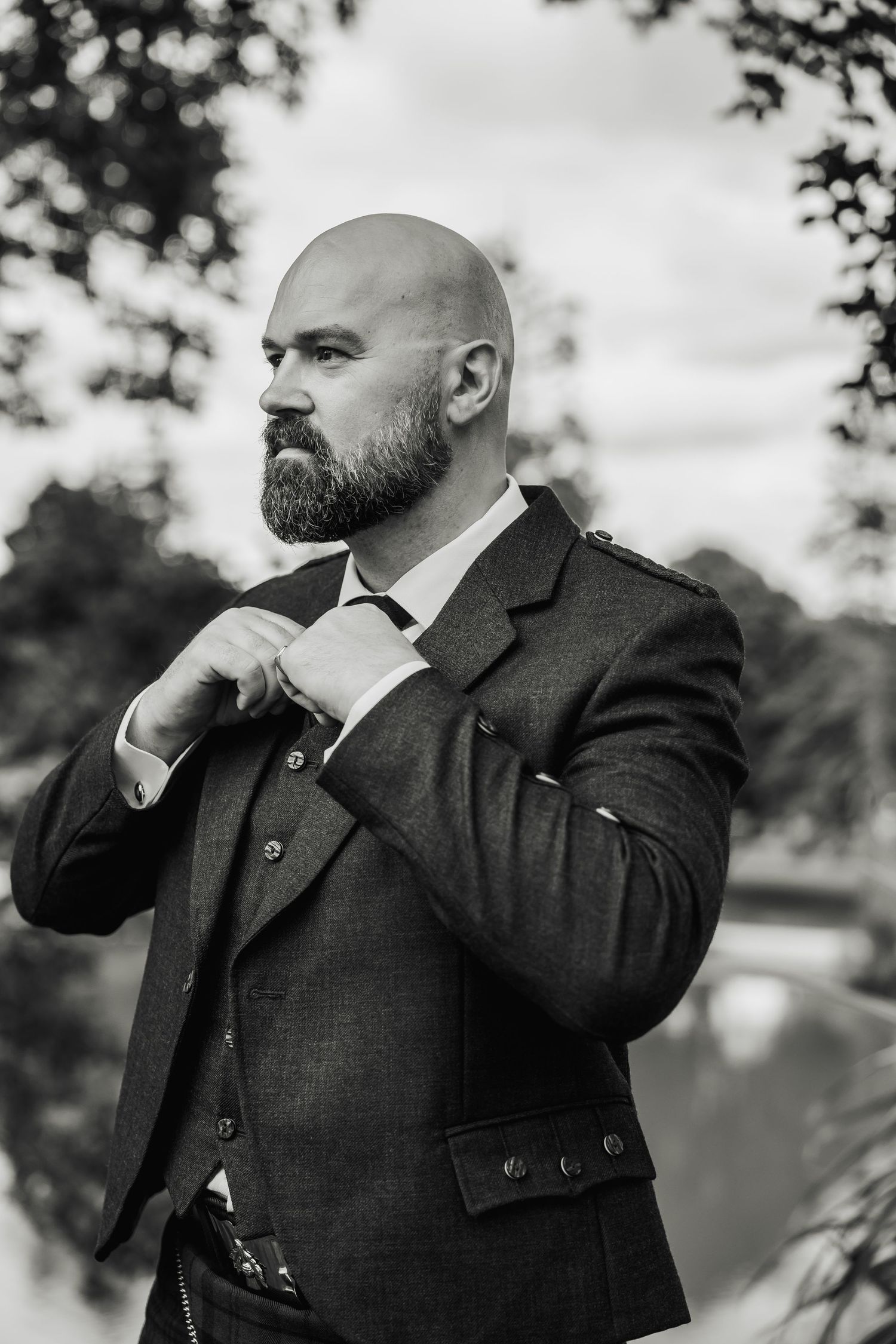 Black and white portrait of a bald bearded groom in a traditional Scottish jacket adjusting his collar outdoors.