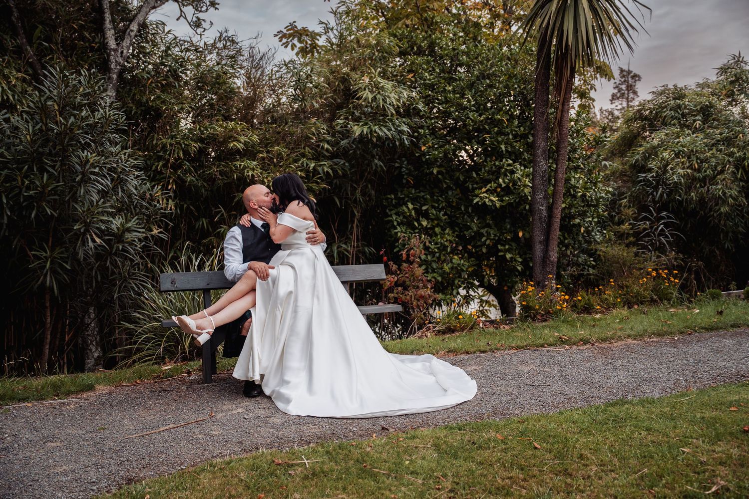 Bride sits on groom's lap sharing a kiss on a garden bench surrounded by lush greenery.