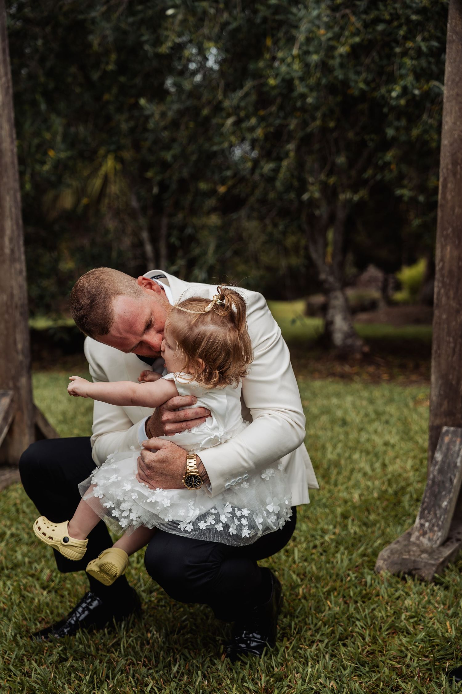 Groom in white jacket tenderly hugging a little girl in a white floral dress outdoors.