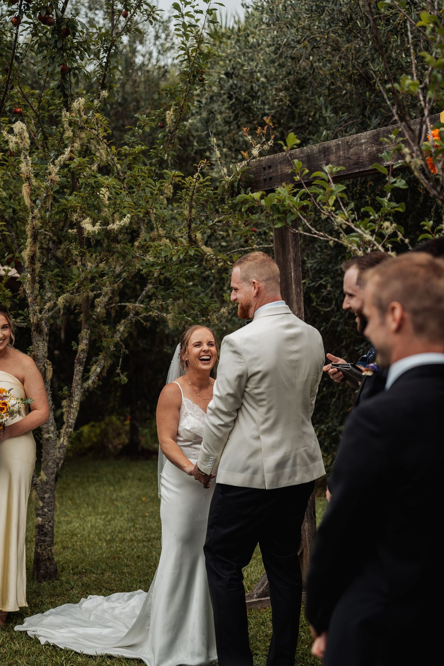 Bride in white gown laughing joyfully with groom in white jacket during outdoor wedding ceremony.