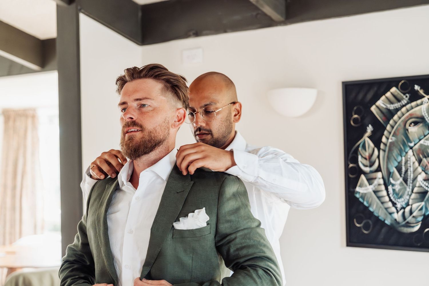 A bald groomsman adjusts the collar of a bearded groom in a green suit before the wedding ceremony.