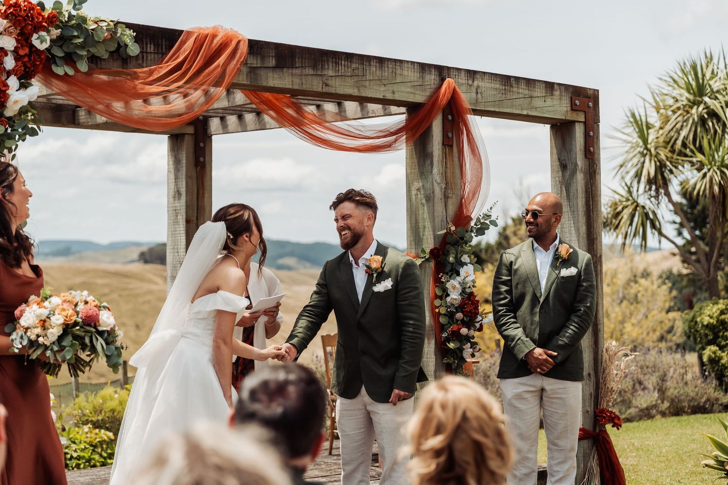 Bride and groom exchange vows under a rustic wooden arch with orange draping and floral decor.