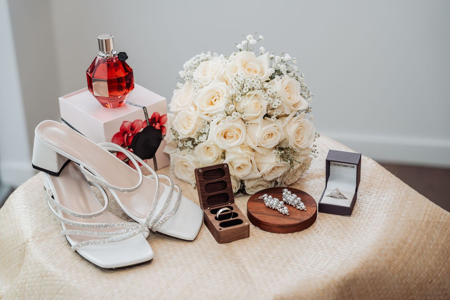 Wedding accessories including white bridal shoes, jewelry boxes, and delicate cream rose bouquets arranged on light fabric.